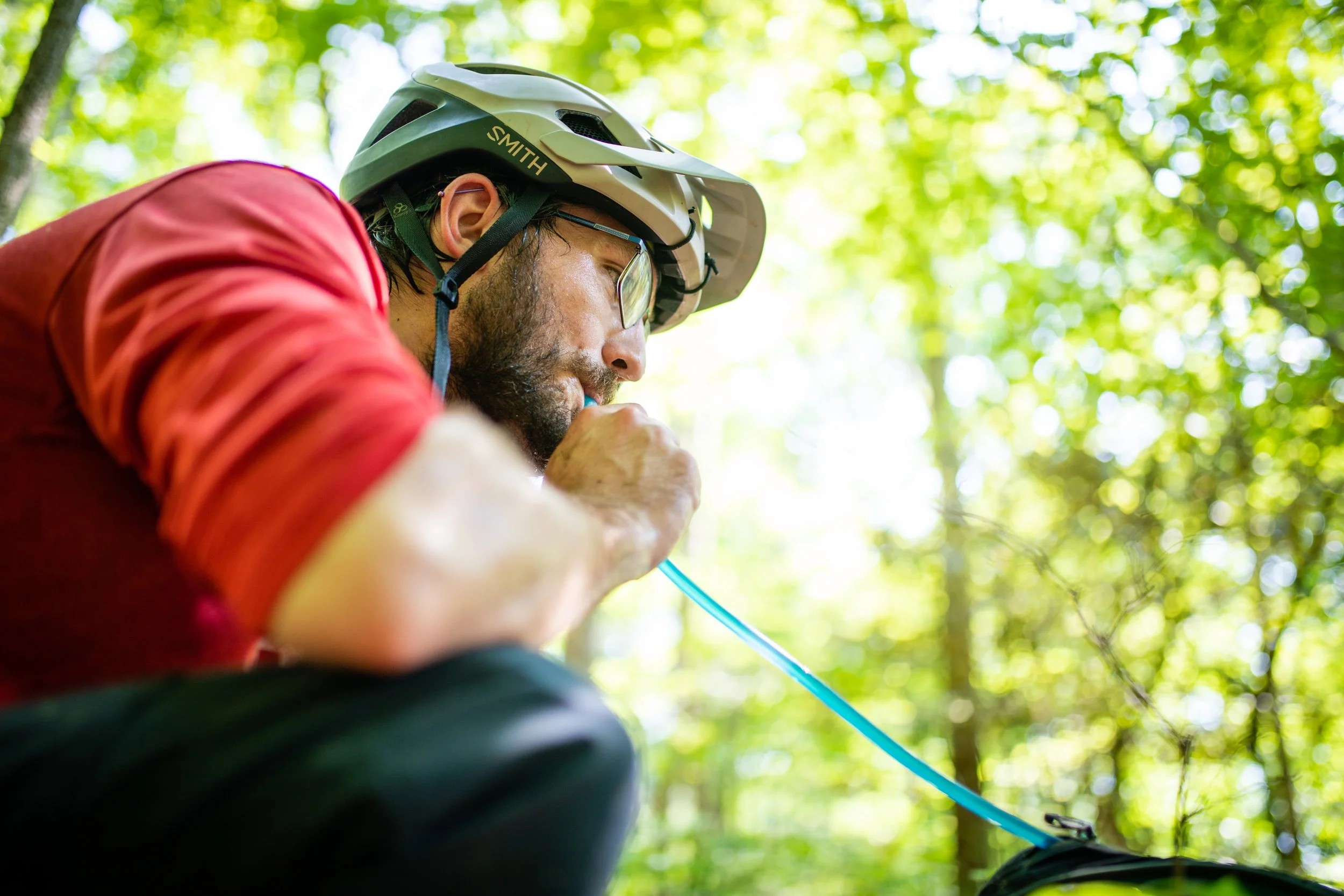 A man with a beard, glasses, and a helmet wearing a red jacket, sitting outdoors in a green forest, drinking from a blue water bottle.