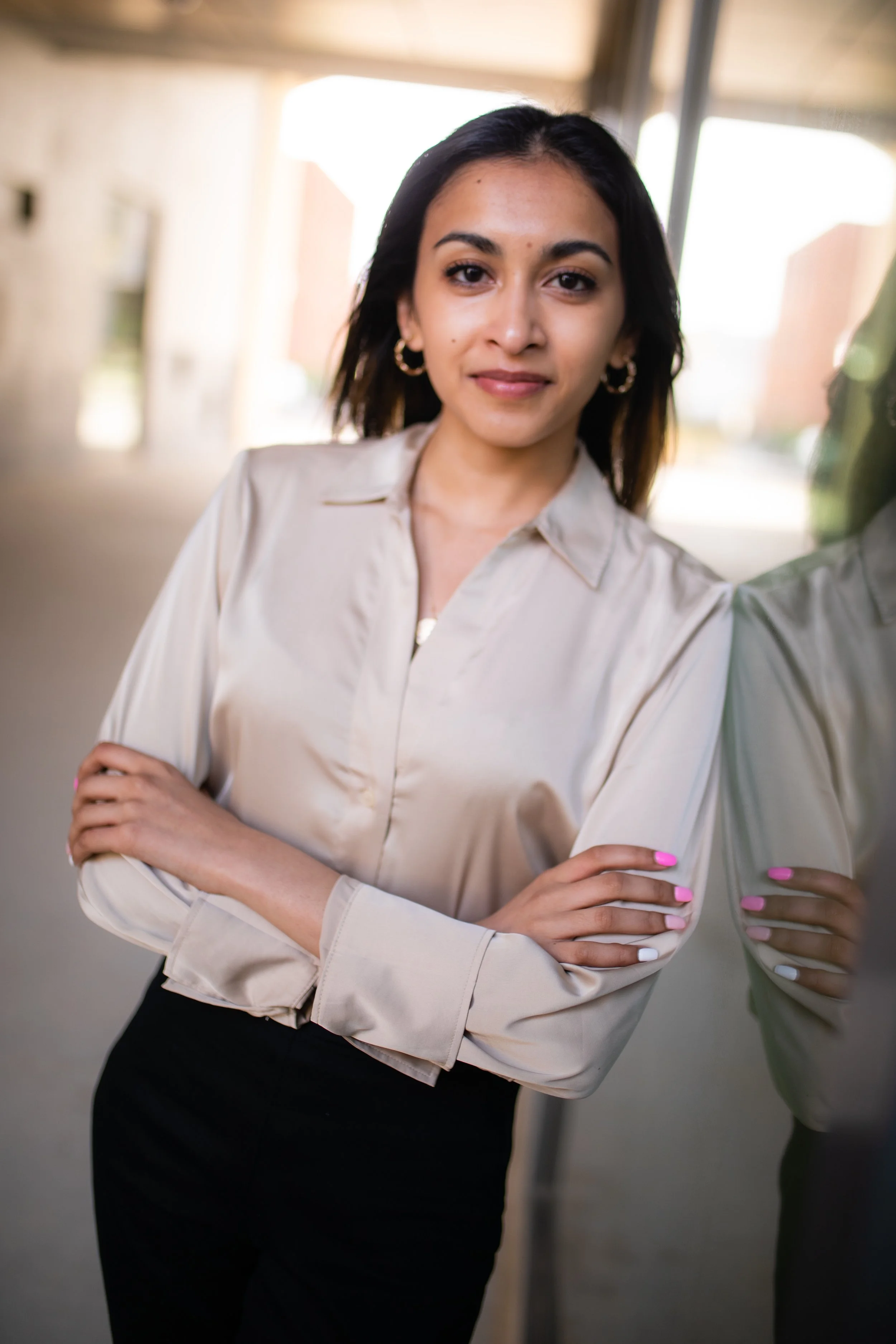 A young woman with dark hair, wearing a beige blouse and black pants, standing indoors near a mirror or glass wall, with her arms crossed and smiling slightly at the camera.