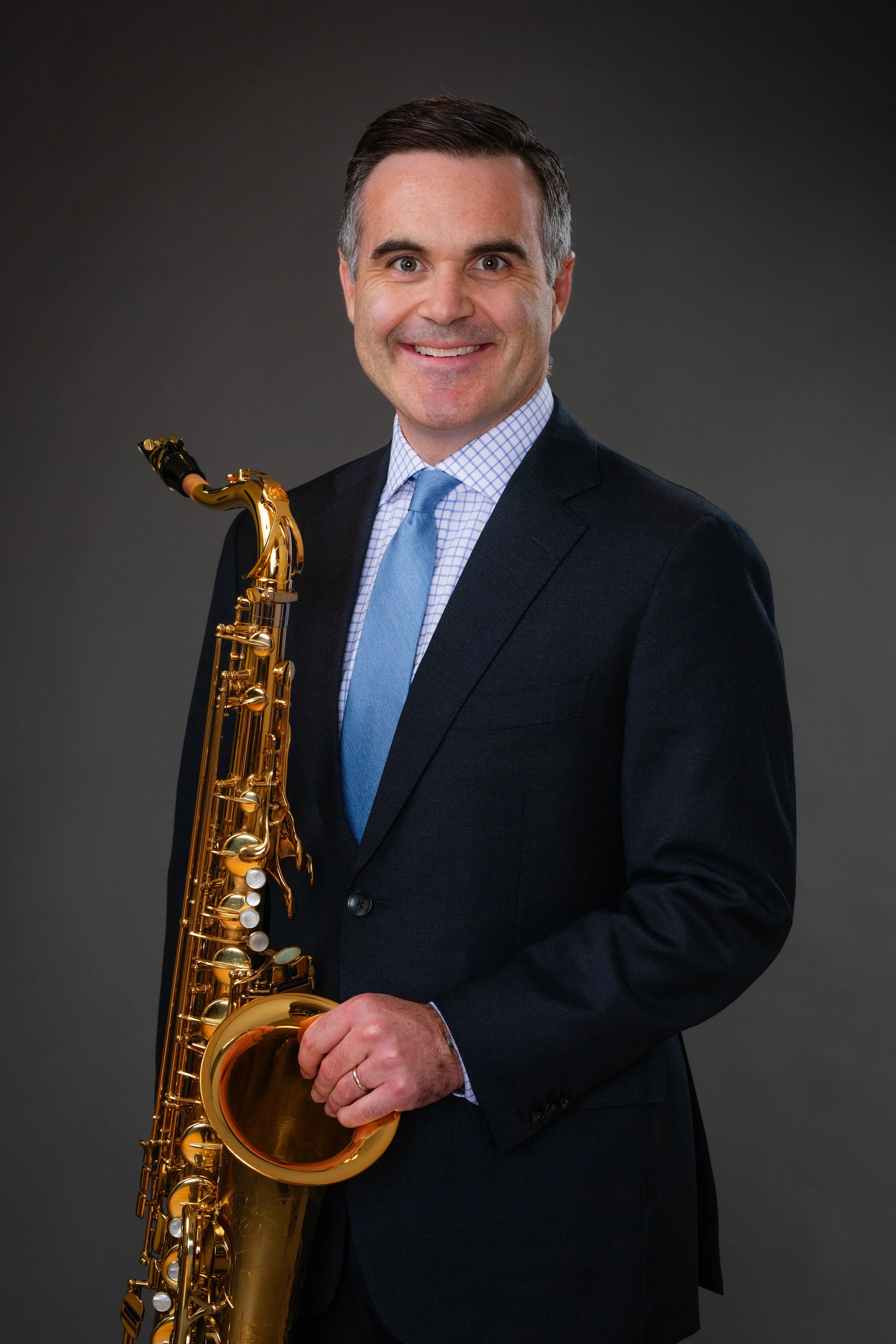 A man in a dark suit with a checked shirt and a blue tie holding a gold saxophone, smiling, in front of a gray background.