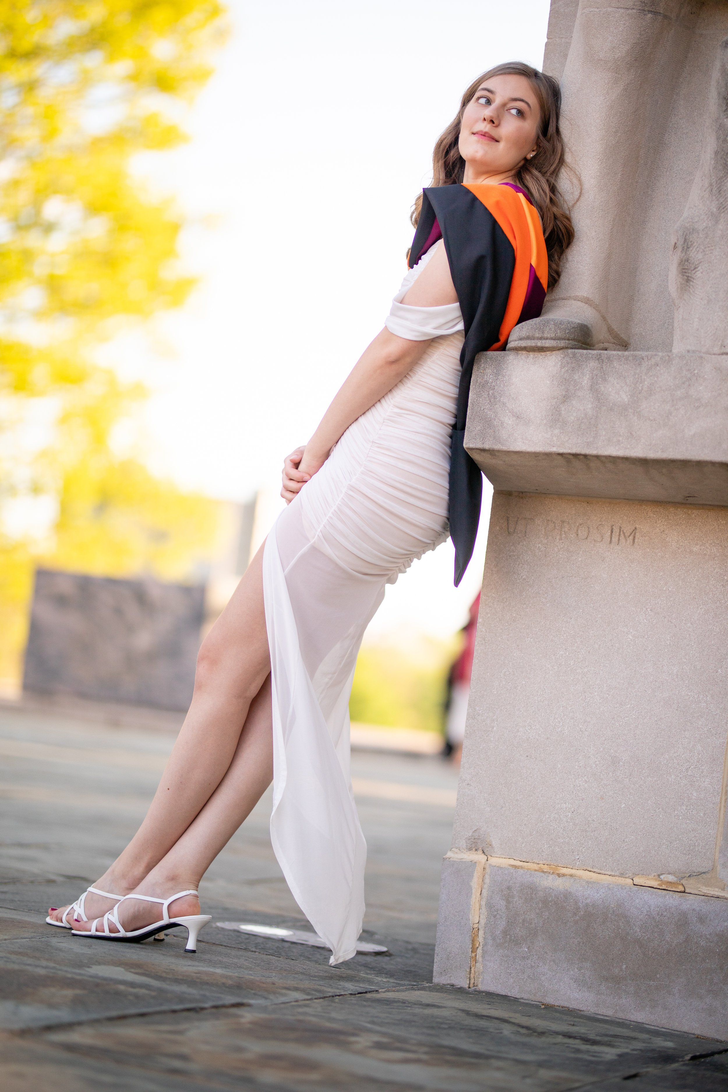 A woman in a white dress with a slit, wearing white high-heeled sandals, leaning against a stone structure outdoors with yellow trees and blue sky in the background.