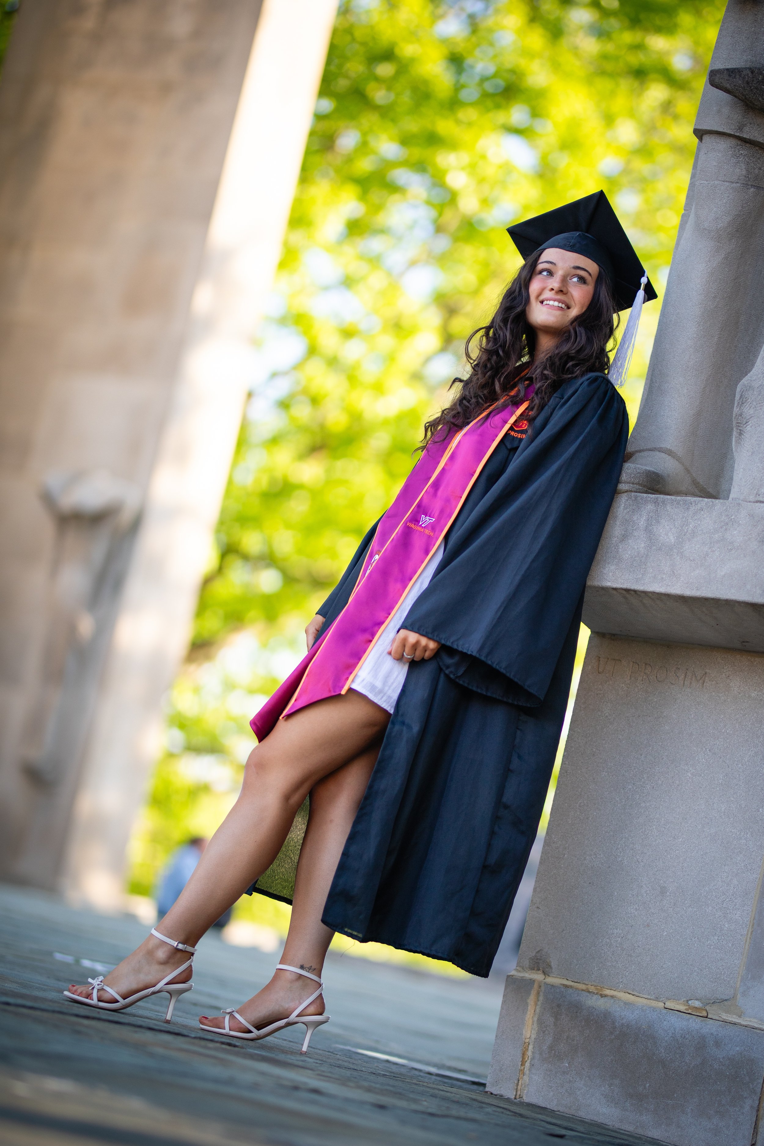 A young woman in graduation gown and cap leaning against a stone sculpture outdoors, smiling, with trees and sunlight in the background.