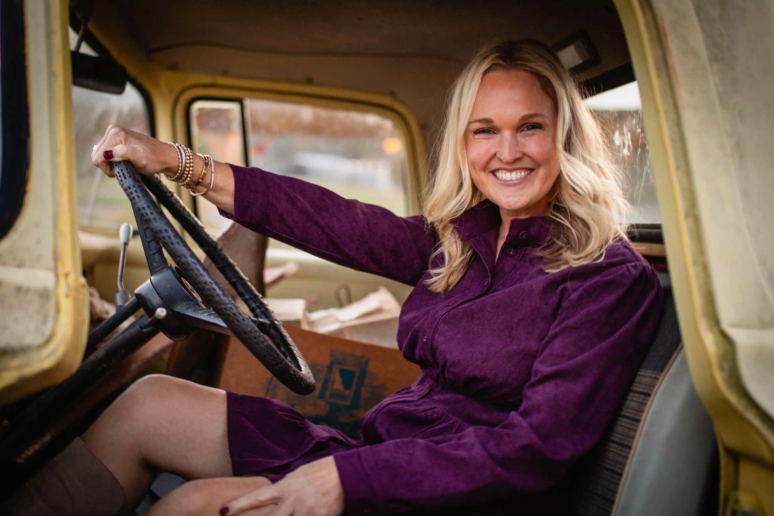 A woman with blonde hair sitting in the driver's seat of a yellow vintage car, smiling and looking at the camera, wearing a purple dress.