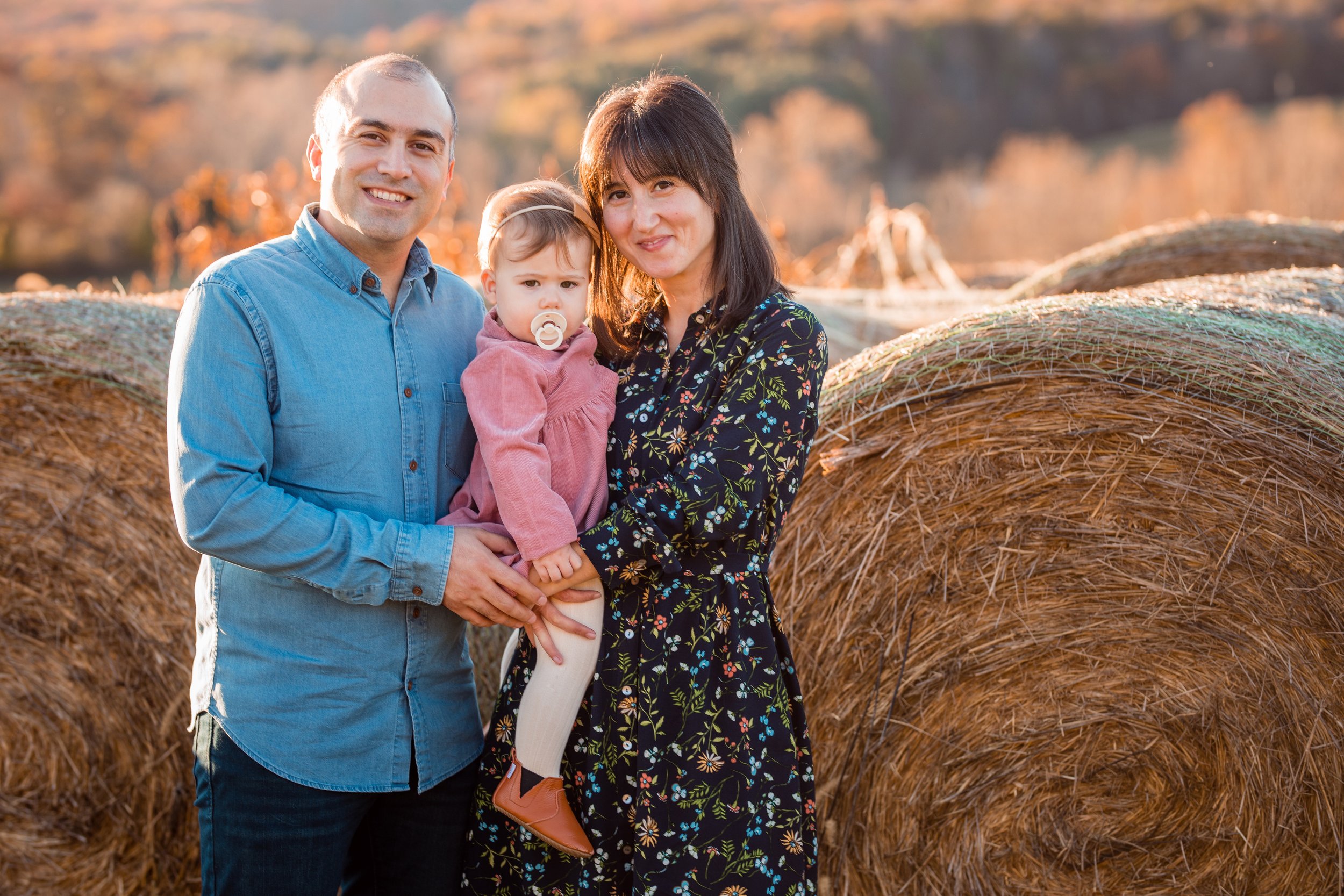A family of three standing outdoors near large hay bales, with autumn trees in the background. The father is on the left, wearing a blue denim shirt. The mother is on the right, wearing a dark floral dress. The child, with a pacifier, is being held b