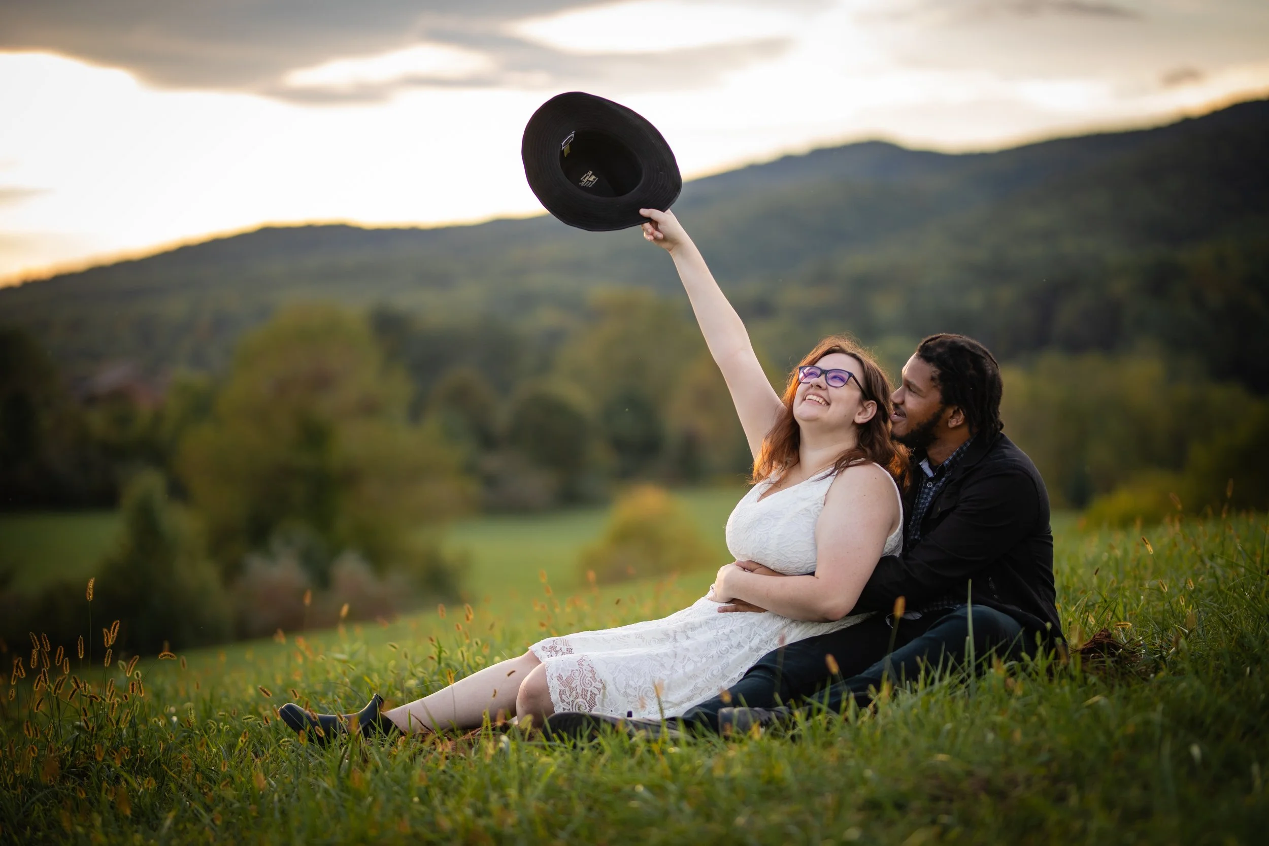A couple sitting on grass in a field, with the woman raising a black hat in the air and smiling, while the man embraces her from behind, during sunset with mountains and trees in the background.