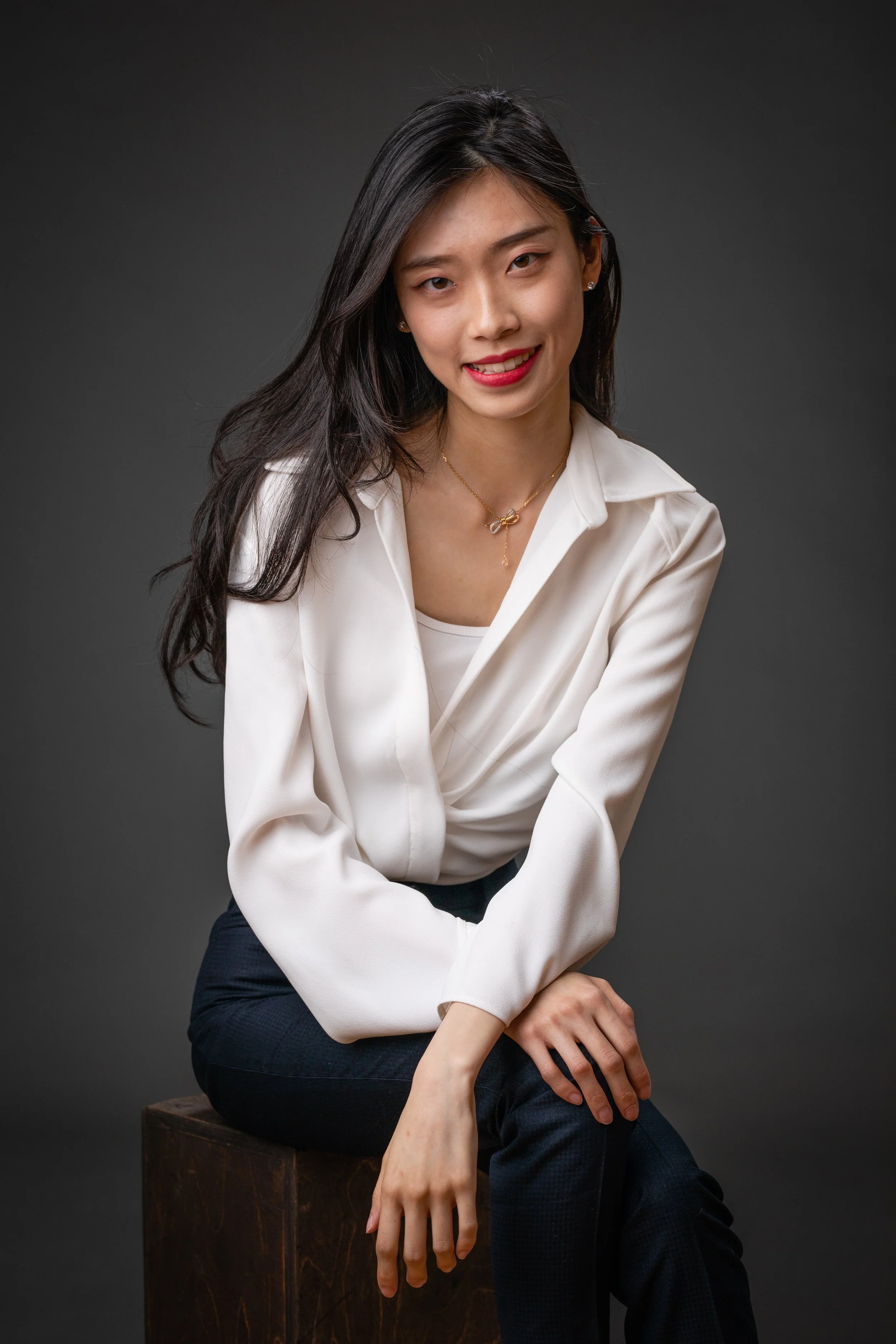 A woman with long dark hair, wearing a white blouse and dark pants, sitting on a wooden block against a plain gray background.