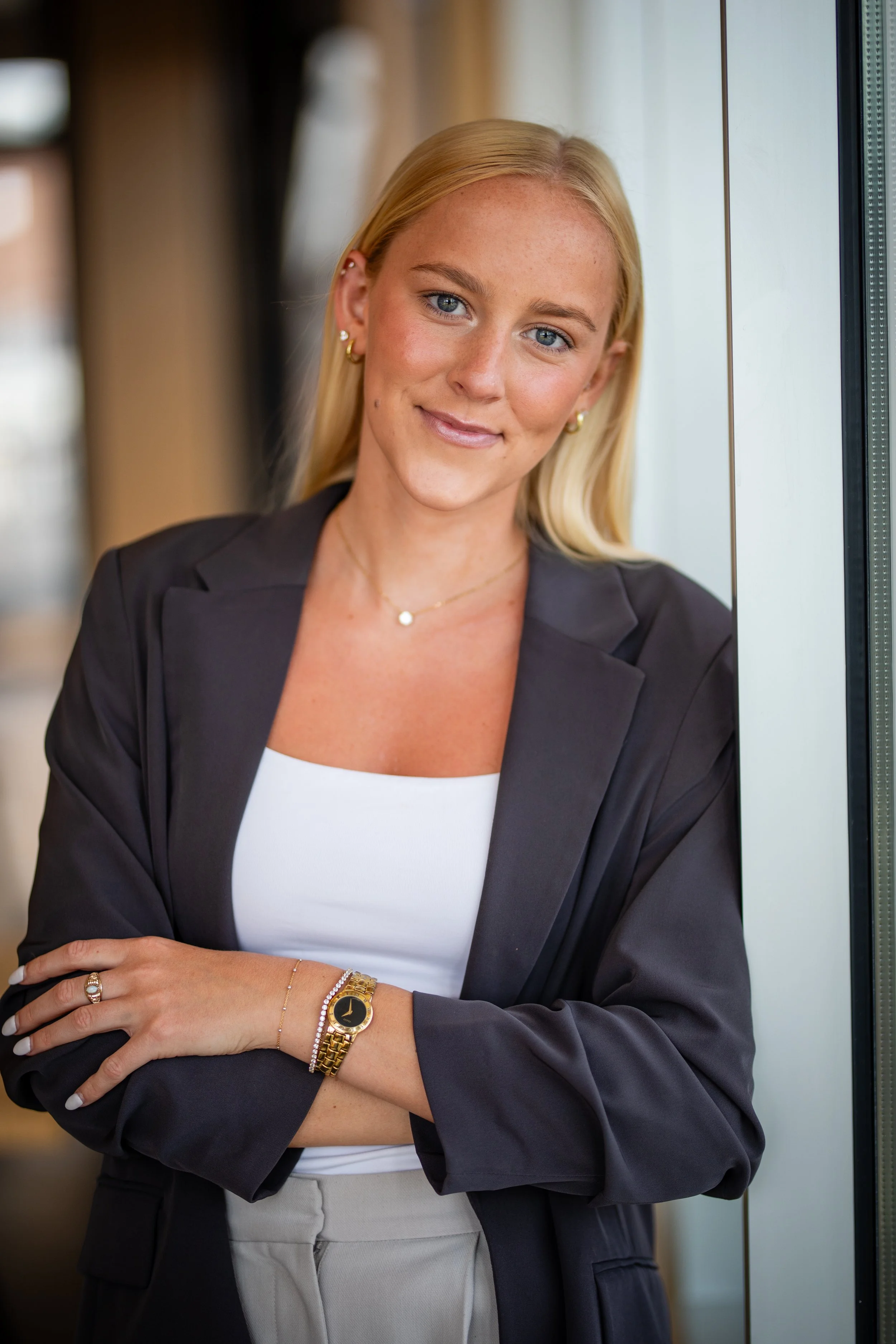 A young woman with blonde hair and blue eyes wearing a black blazer, white top, and beige pants, standing with arms crossed near a window.