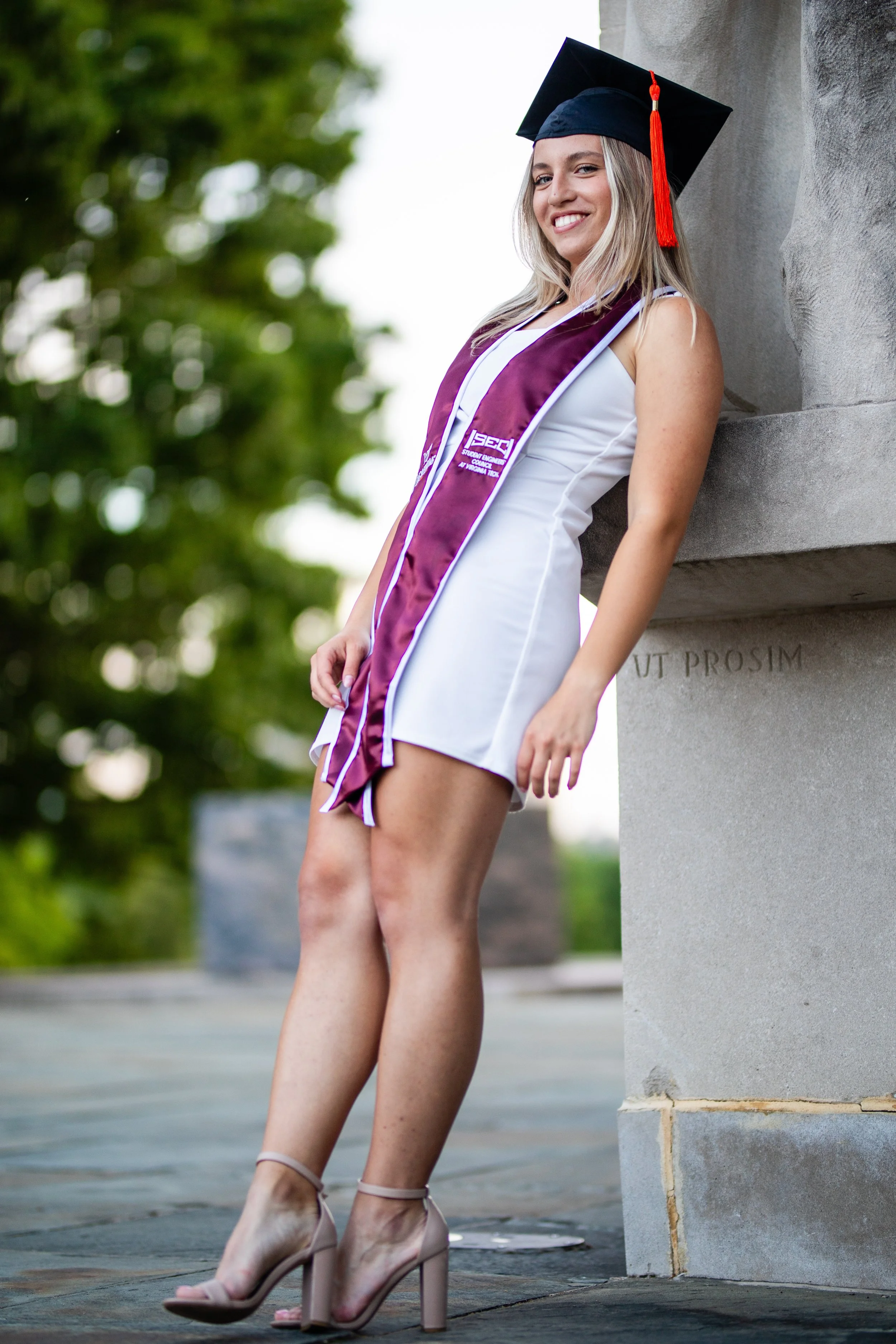 Female graduate in a white dress, wearing a black graduation cap with a red tassel and a purple stole, standing outdoors near a stone monument, smiling.
