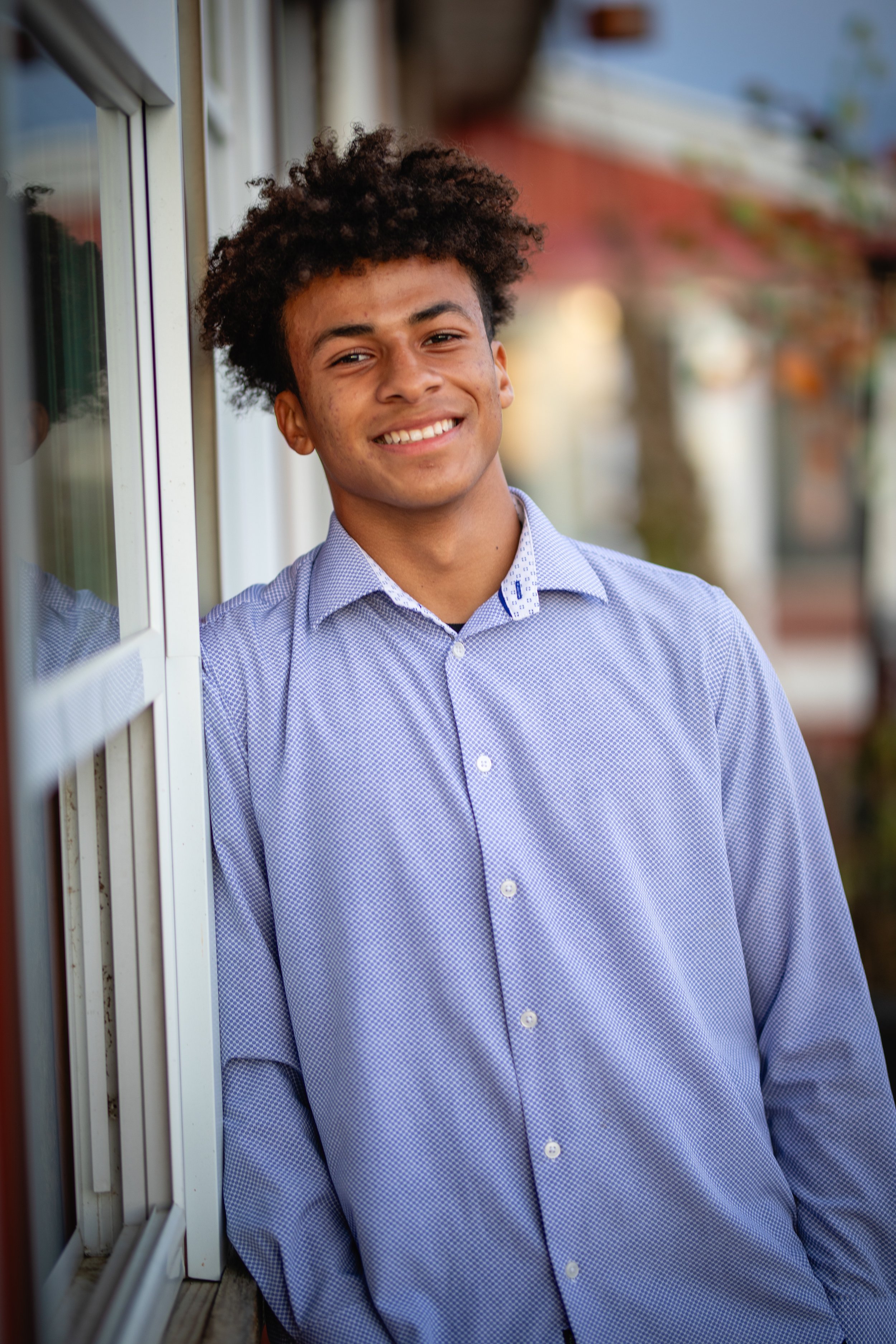 A young man with curly hair smiling and leaning against a window outdoors with trees in the background.