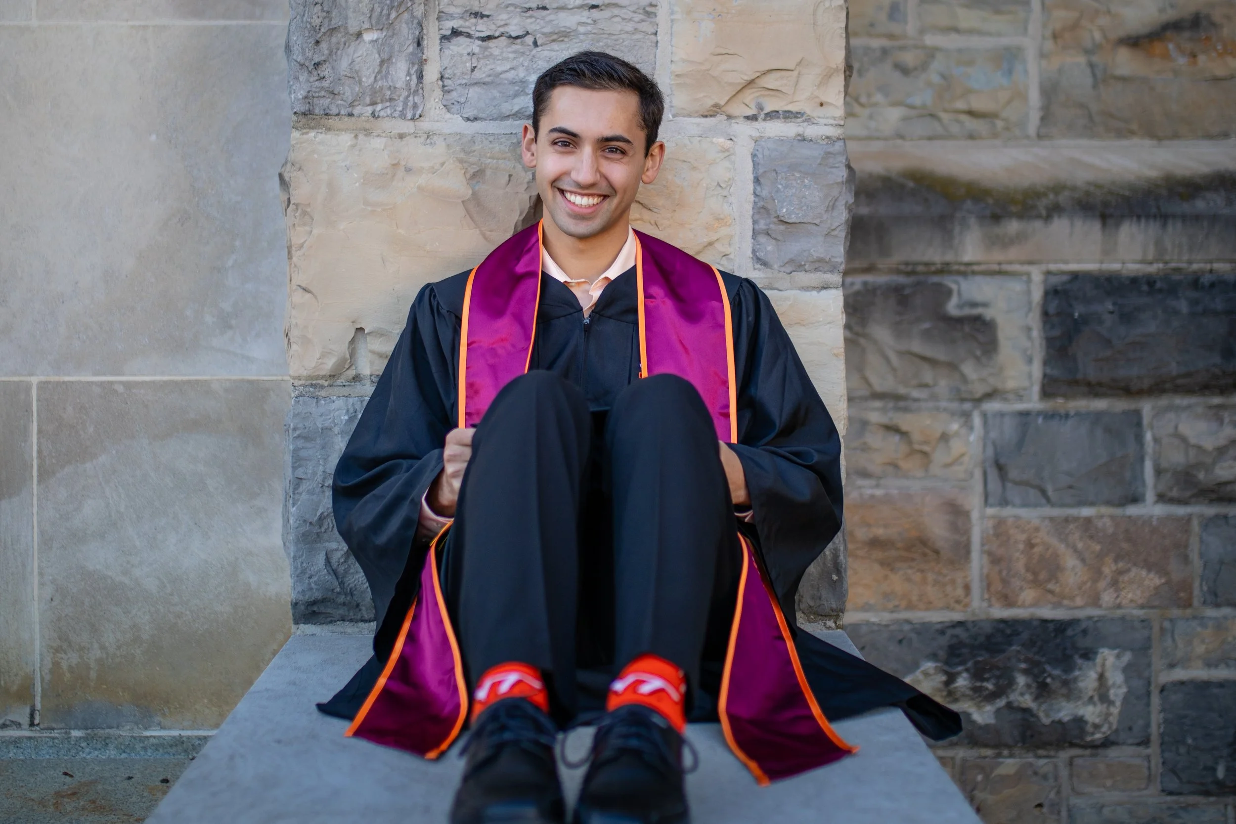 Young man in a graduation cap and gown sitting on a stone ledge, smiling, with a stone wall background.