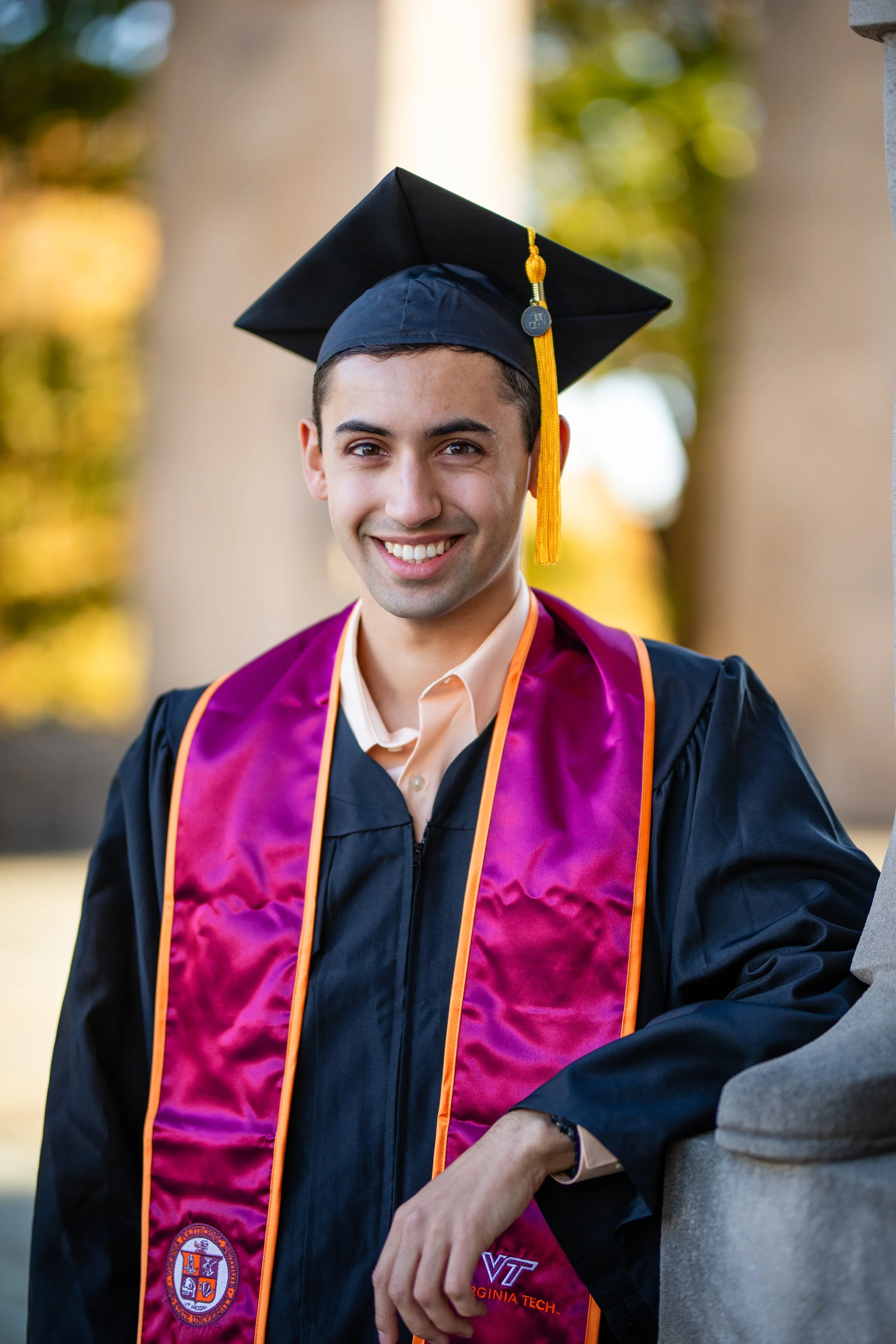 A young man in a graduation cap and gown with a purple and gold stole, smiling outdoors during daytime.