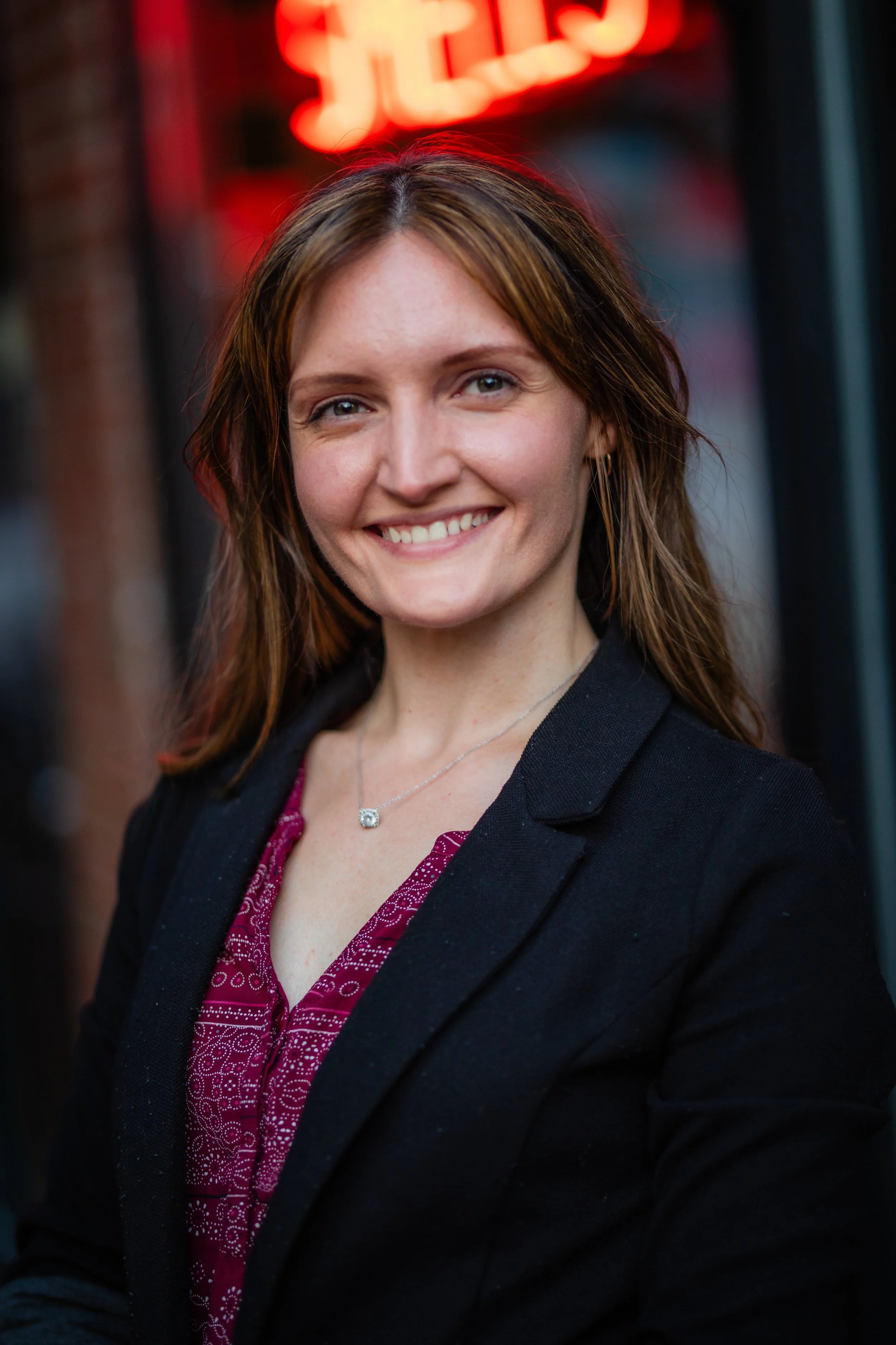A young woman with shoulder-length brown hair, smiling, standing outdoors at dusk or evening, wearing a black blazer over a patterned maroon blouse, with a necklace, in front of a brick building with a red neon sign.