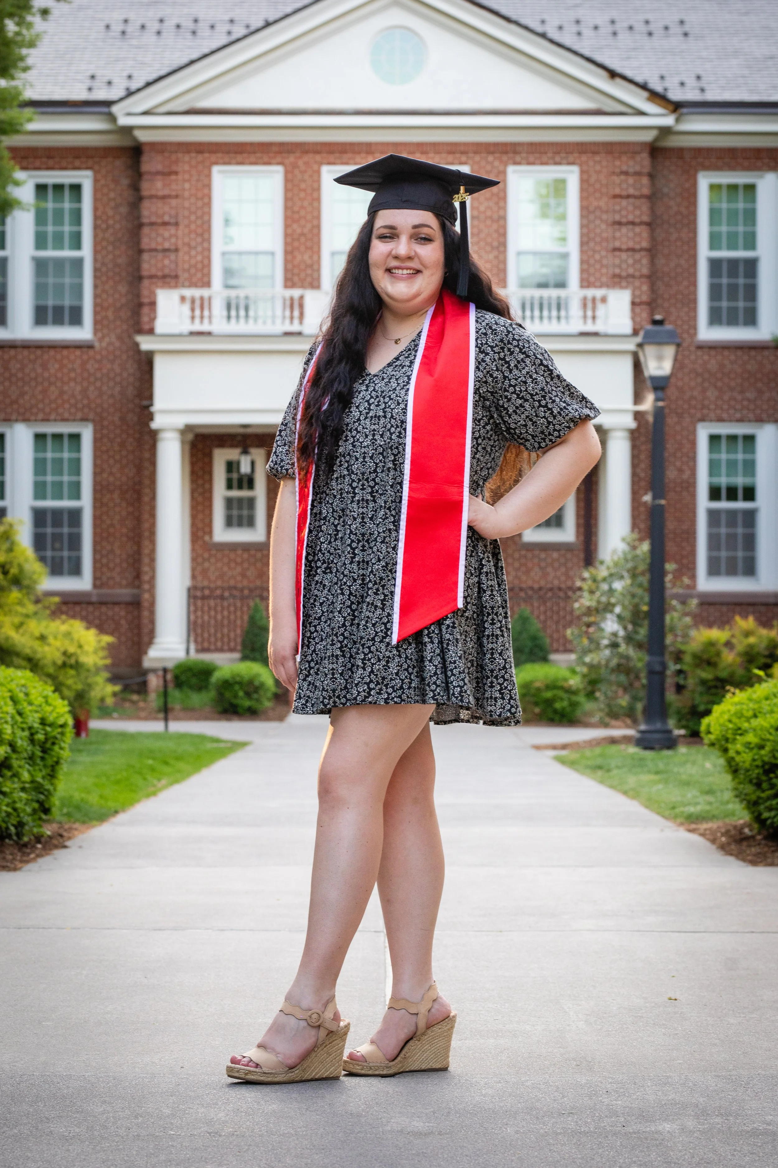 A young woman wearing a black mortarboard and red stole, smiling, outdoors in front of a large brick house, posing with her hand on her hip.