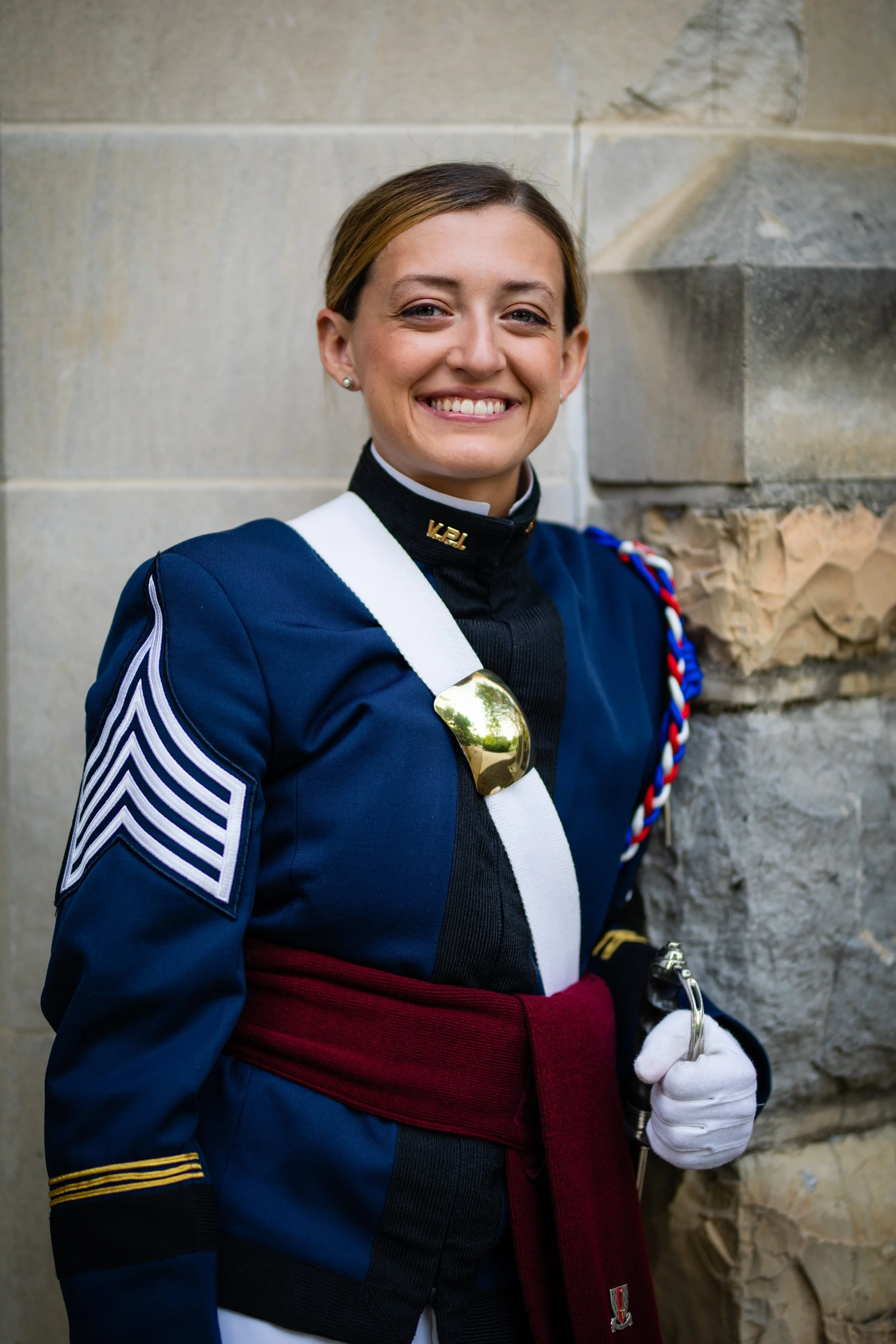 A woman in a military uniform smiling and holding a baton, standing against a stone wall.