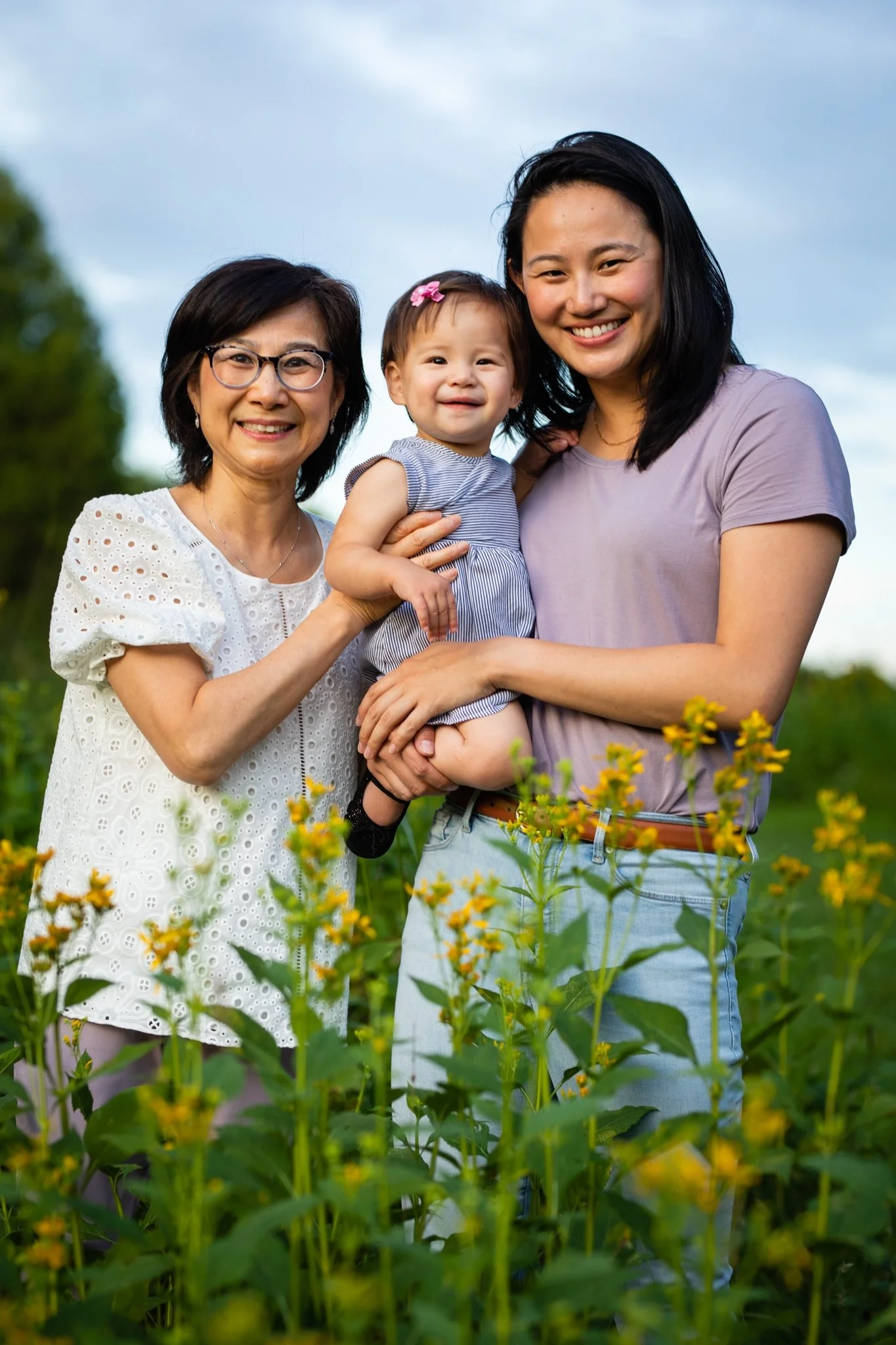 Three women, one older and two younger, holding a young girl in a field of yellow flowers outdoors under a blue sky.