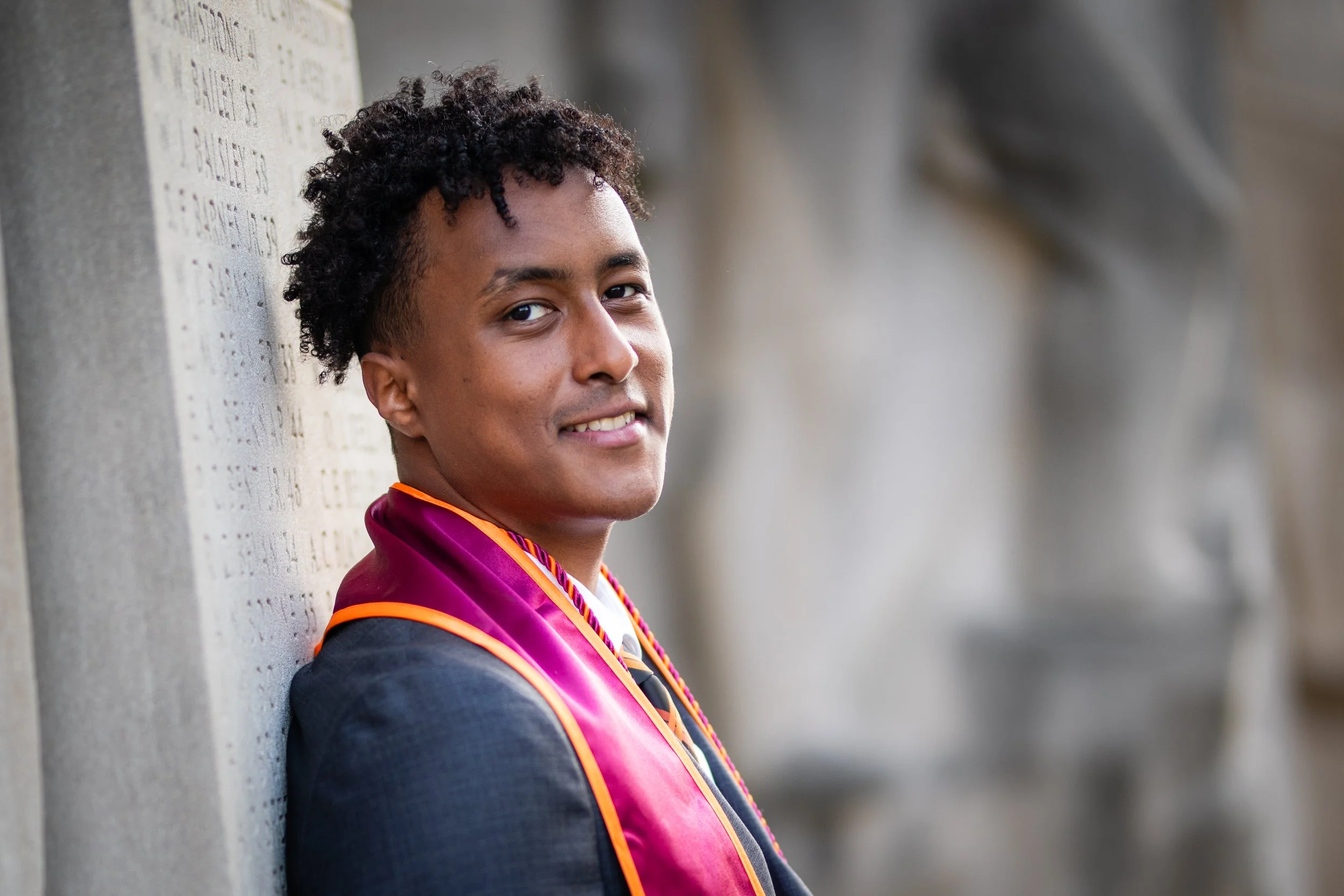 Young man in graduation gown and hood, smiling, leaning against a wall with engraved text, outdoors.