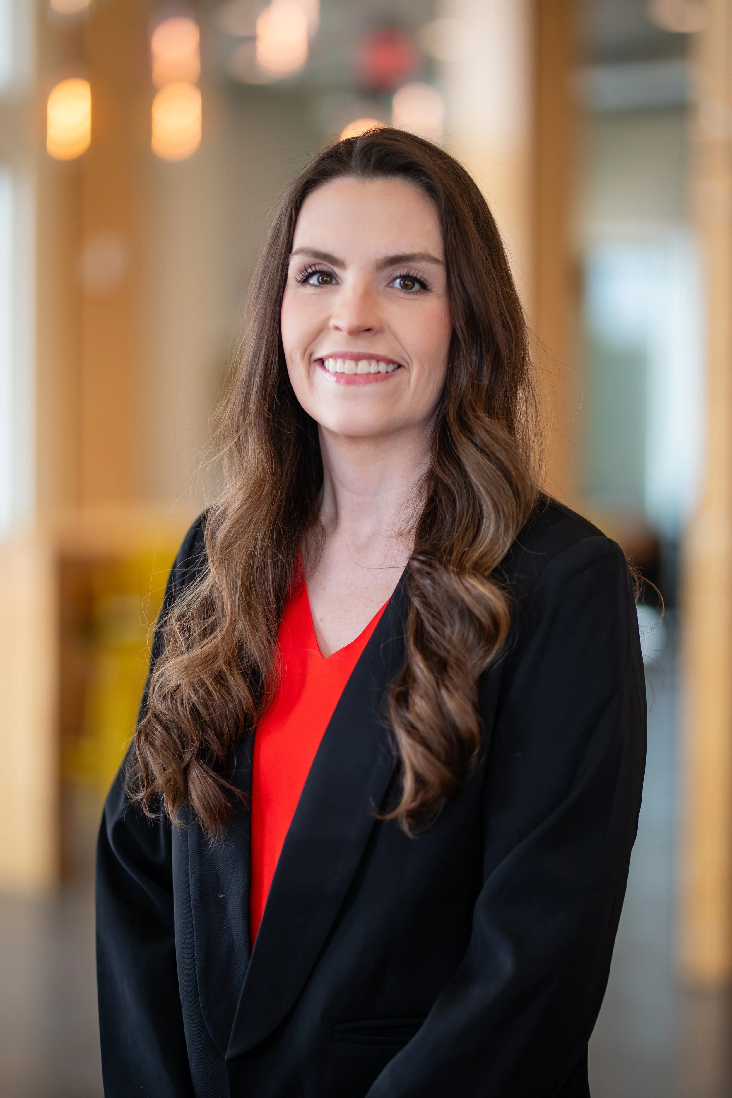 Portrait of a smiling woman with long brown hair, wearing a black blazer and a red blouse, standing inside a modern office or restaurant with warm lighting.