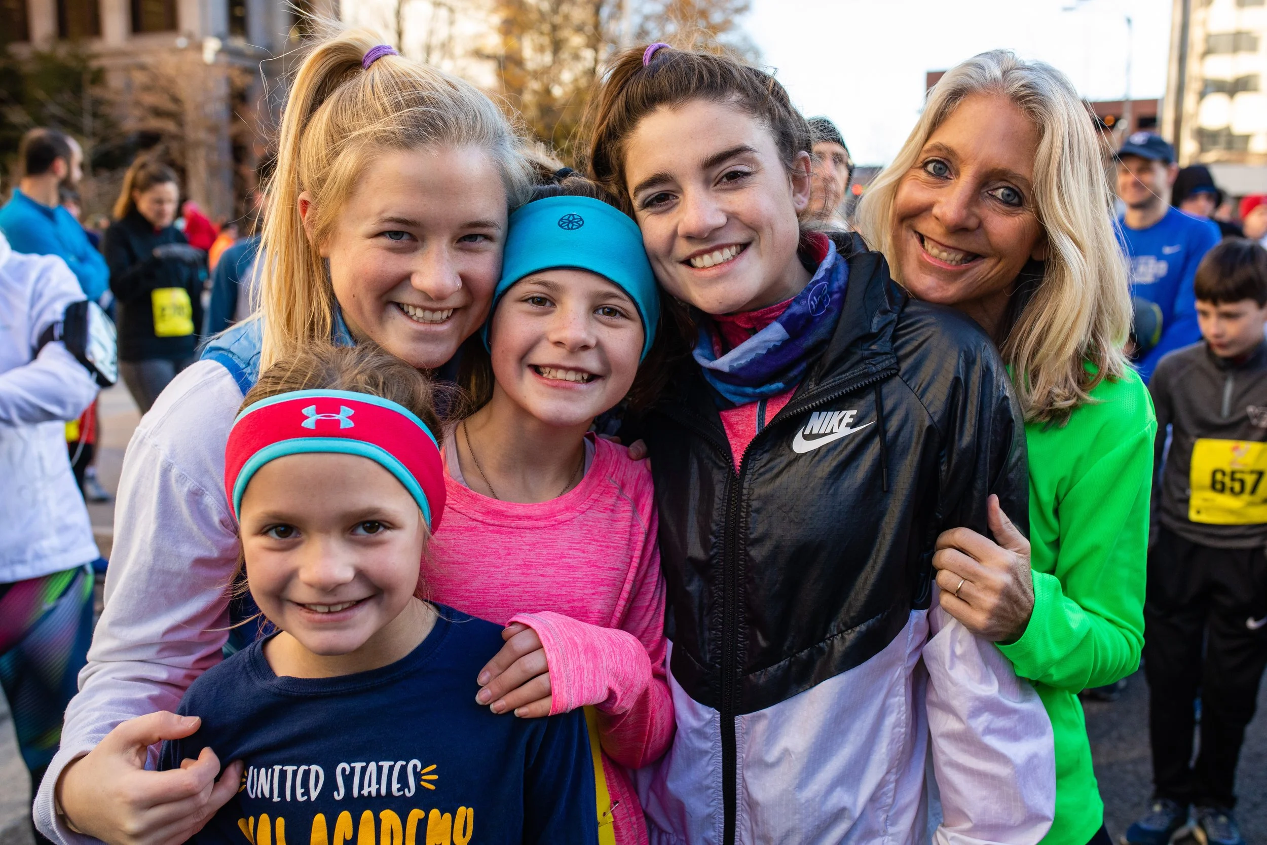 A group of five smiling women and girls, including a young girl in a navy blue shirt and bright pink headband, standing outdoors at a race or event, with multiple people in the background.