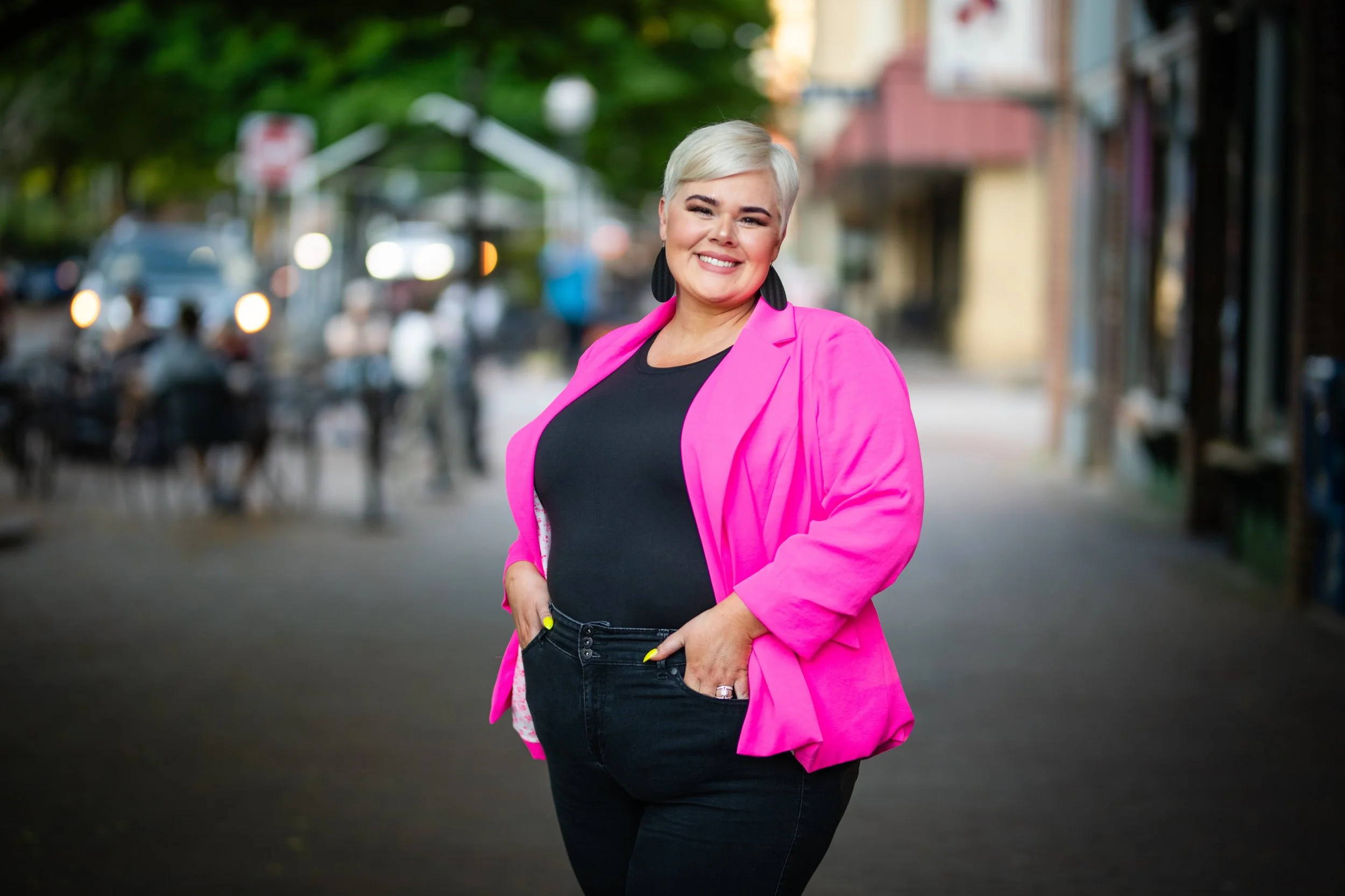 Smiling woman with short blonde hair wearing a pink blazer and black top, standing on an urban street.