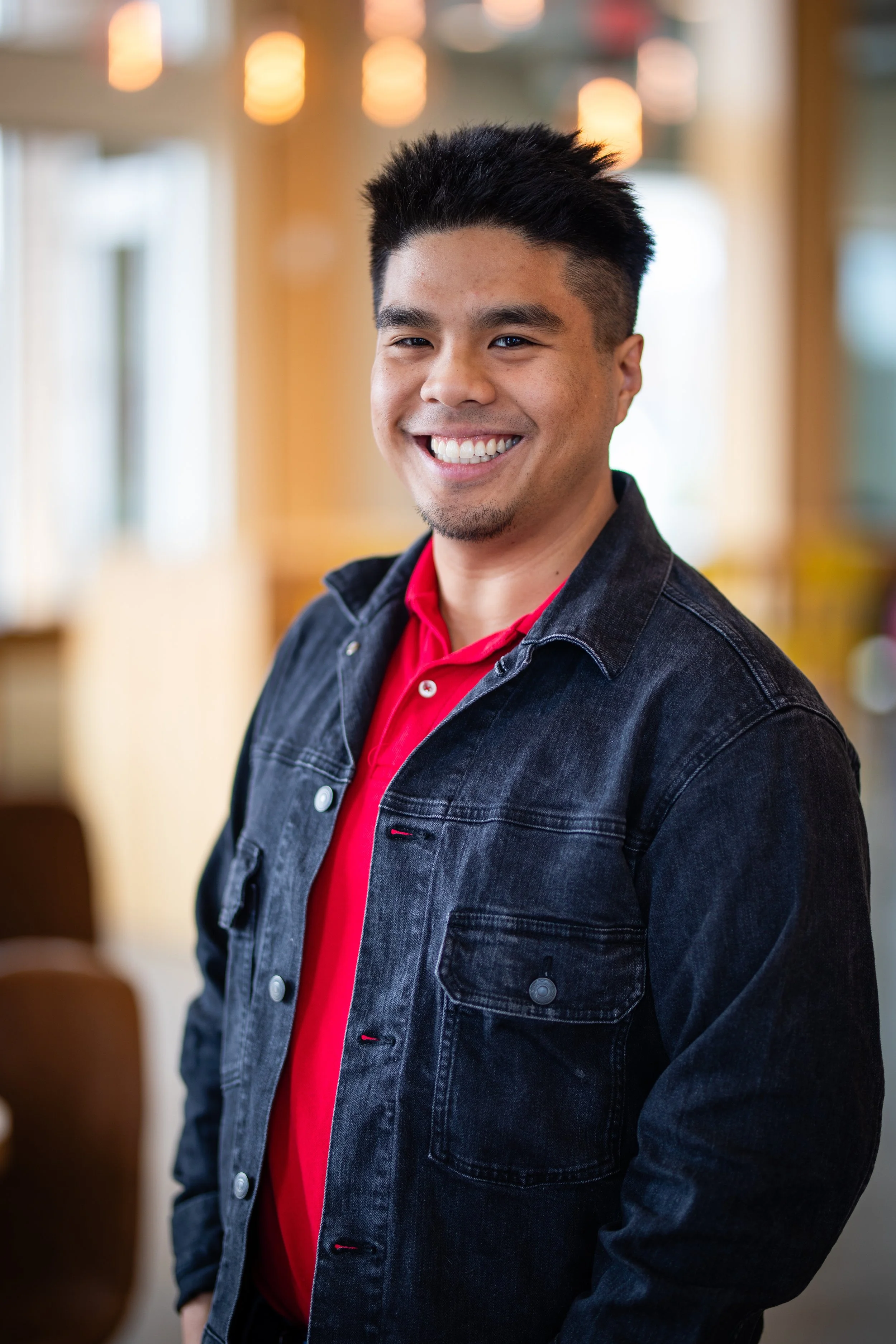 A young man smiling, wearing a red shirt and black denim jacket, standing inside a warmly lit room.