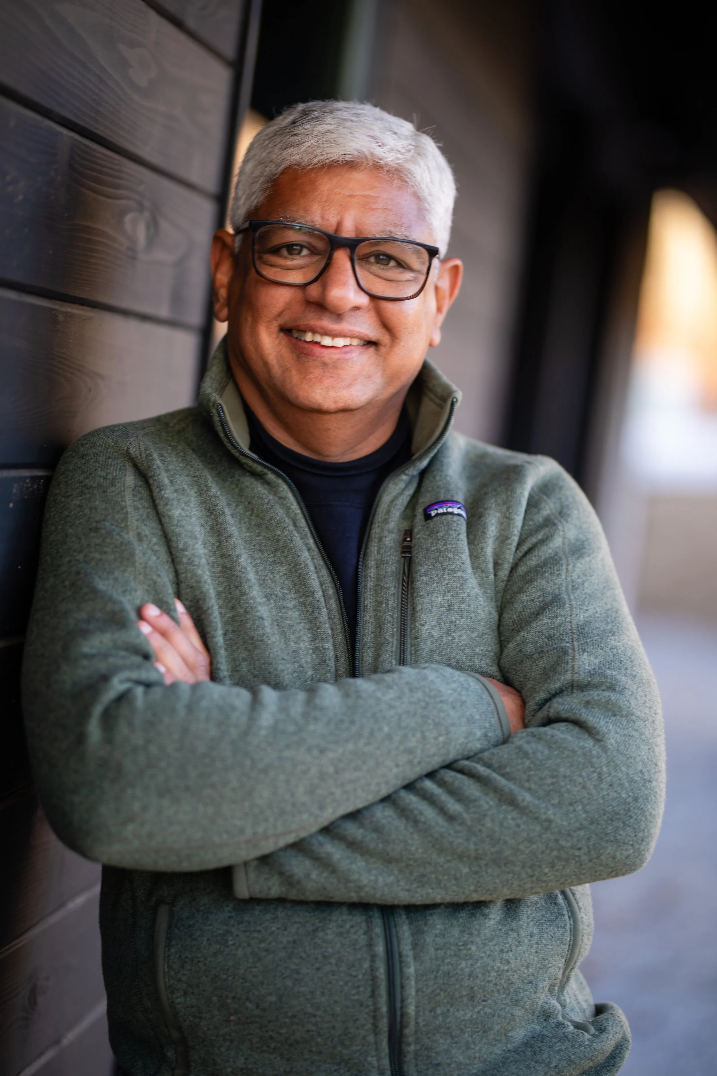 A middle-aged man with gray hair wearing glasses and a green Patagonia fleece jacket, smiling and standing with arms crossed against a wooden wall outdoors.