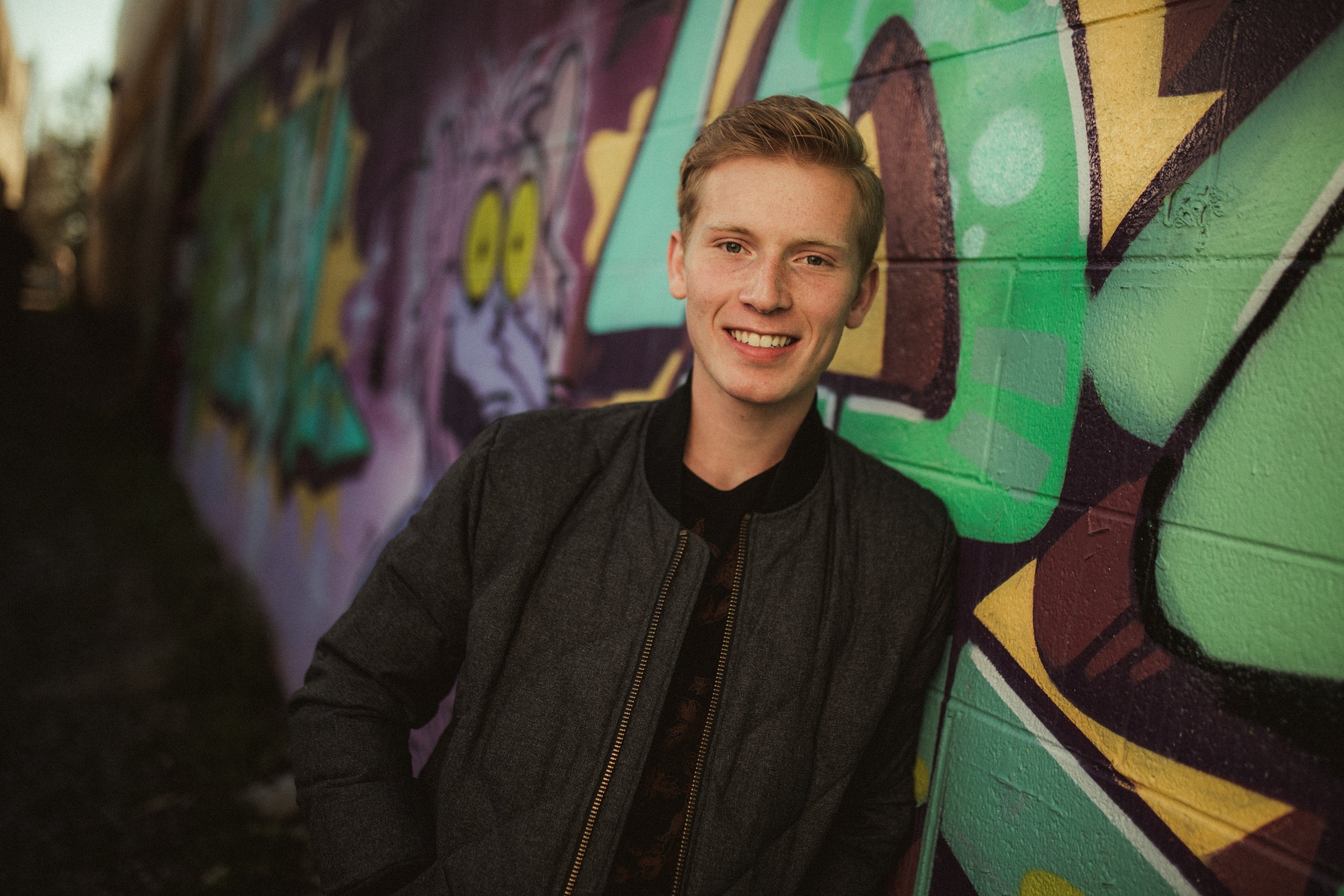 A young man smiling and leaning against a colorful graffiti mural on a concrete wall.