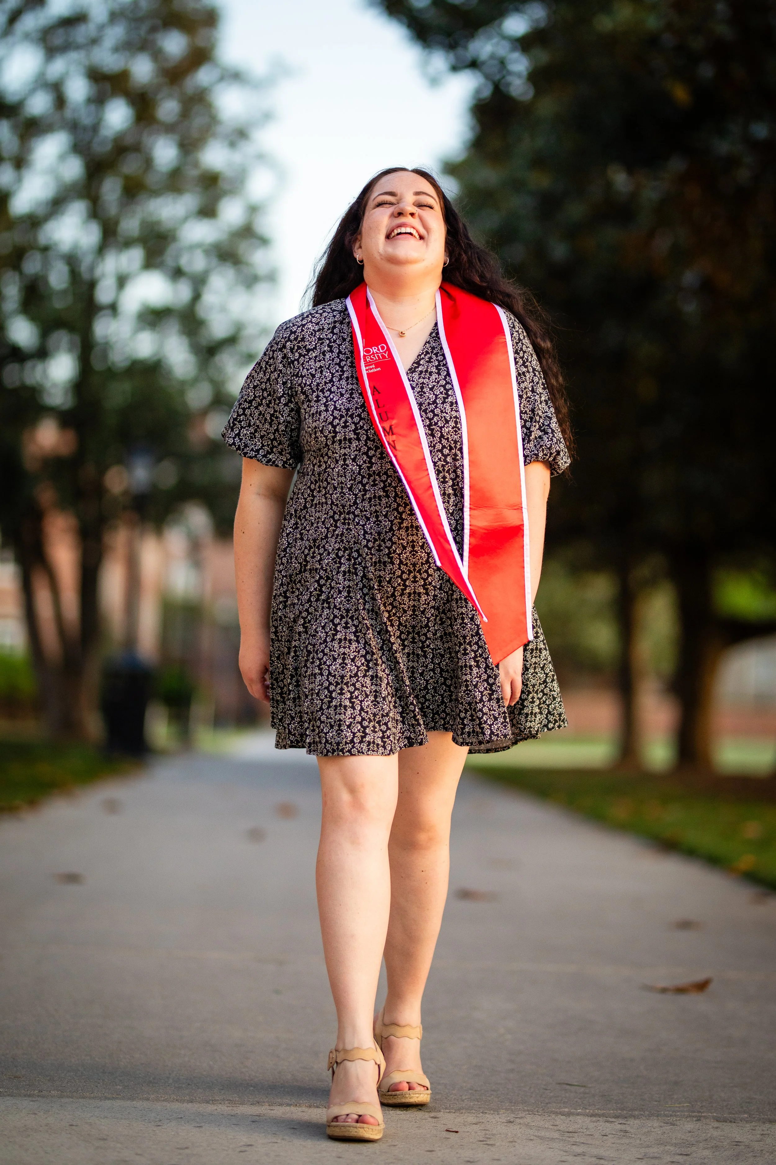 A woman in a black floral dress is smiling and walking outdoors, wearing a red graduation stole and a congratulatory sash.