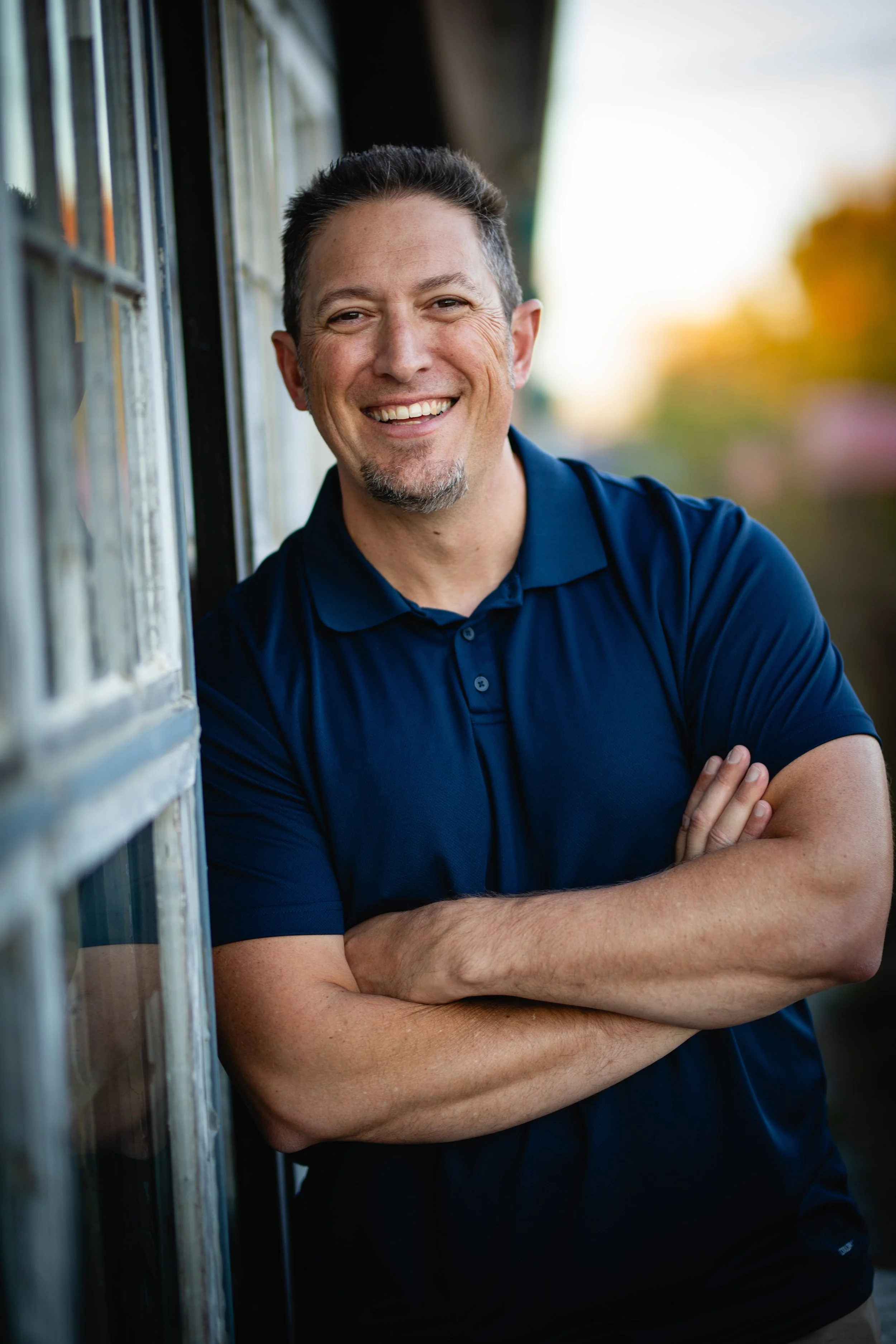 A man with short dark hair, a goatee, and a bright smile, standing outdoors with arms crossed, wearing a dark blue polo shirt, leaning against a wooden structure with a blurred autumnal background.