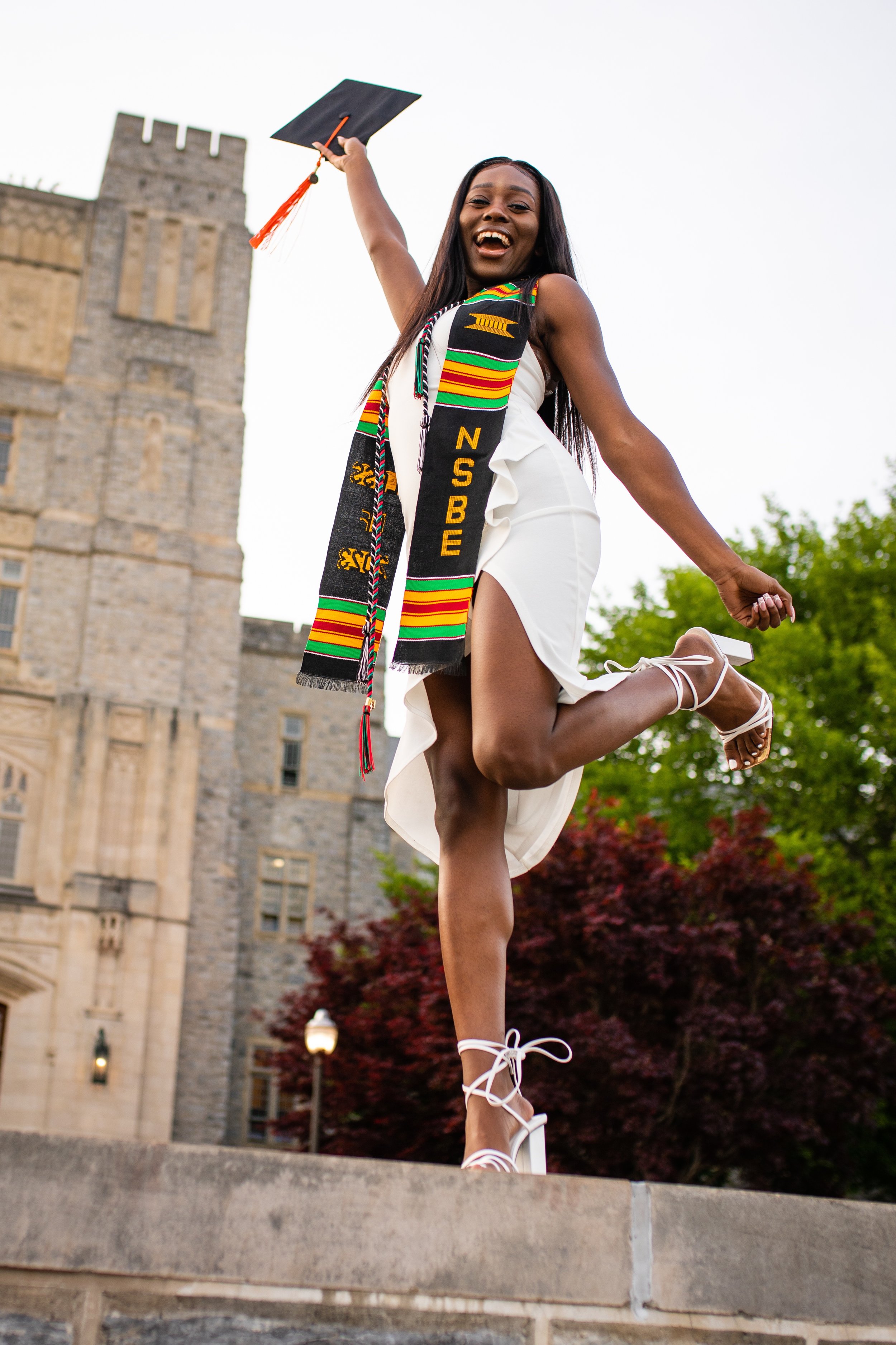 A young woman in a white dress and white high heels celebrating her graduation outdoors, holding a cap in one hand and jumping with joy.