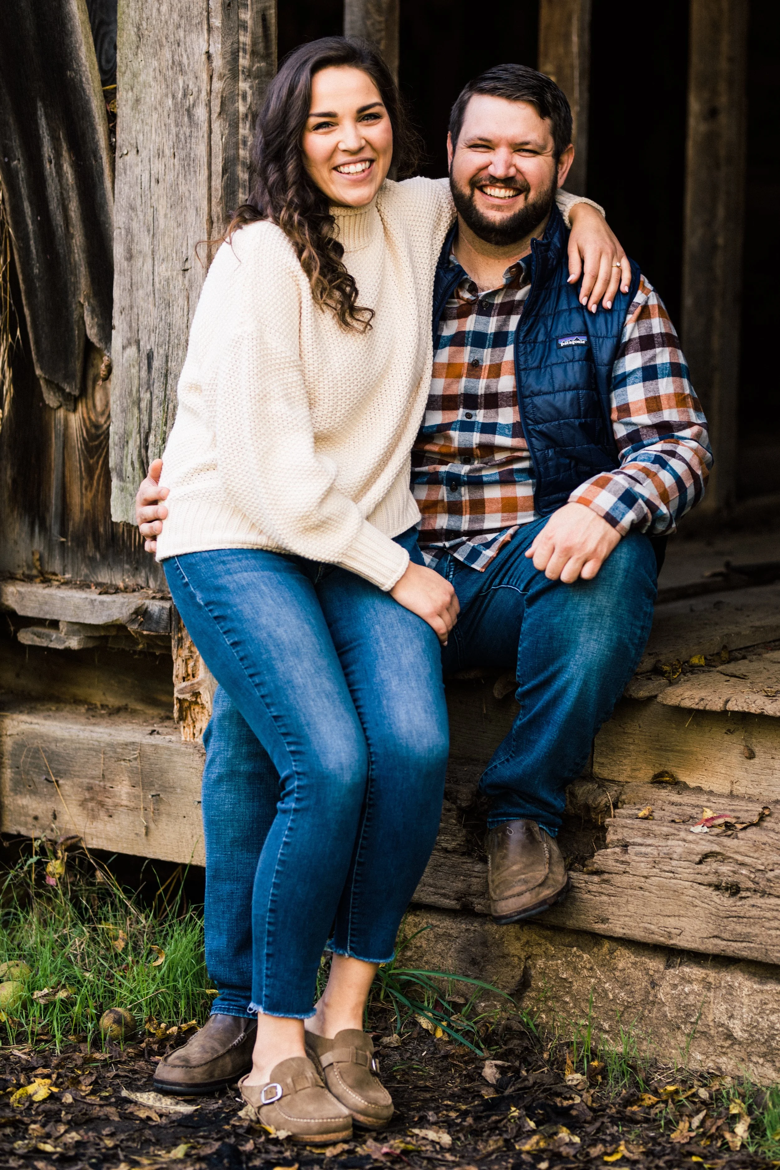 A smiling couple sitting outdoors, leaning against a rustic wooden structure, dressed in casual outdoor clothing.