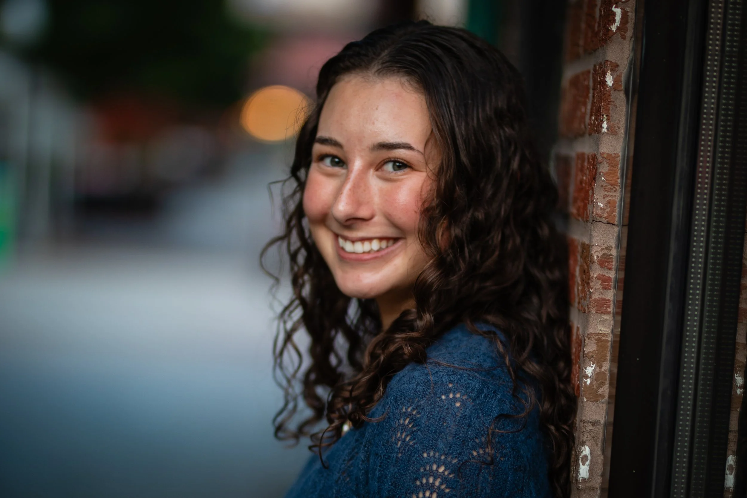 A young woman with long, curly brown hair smiling and leaning against a brick wall outdoors at dusk.