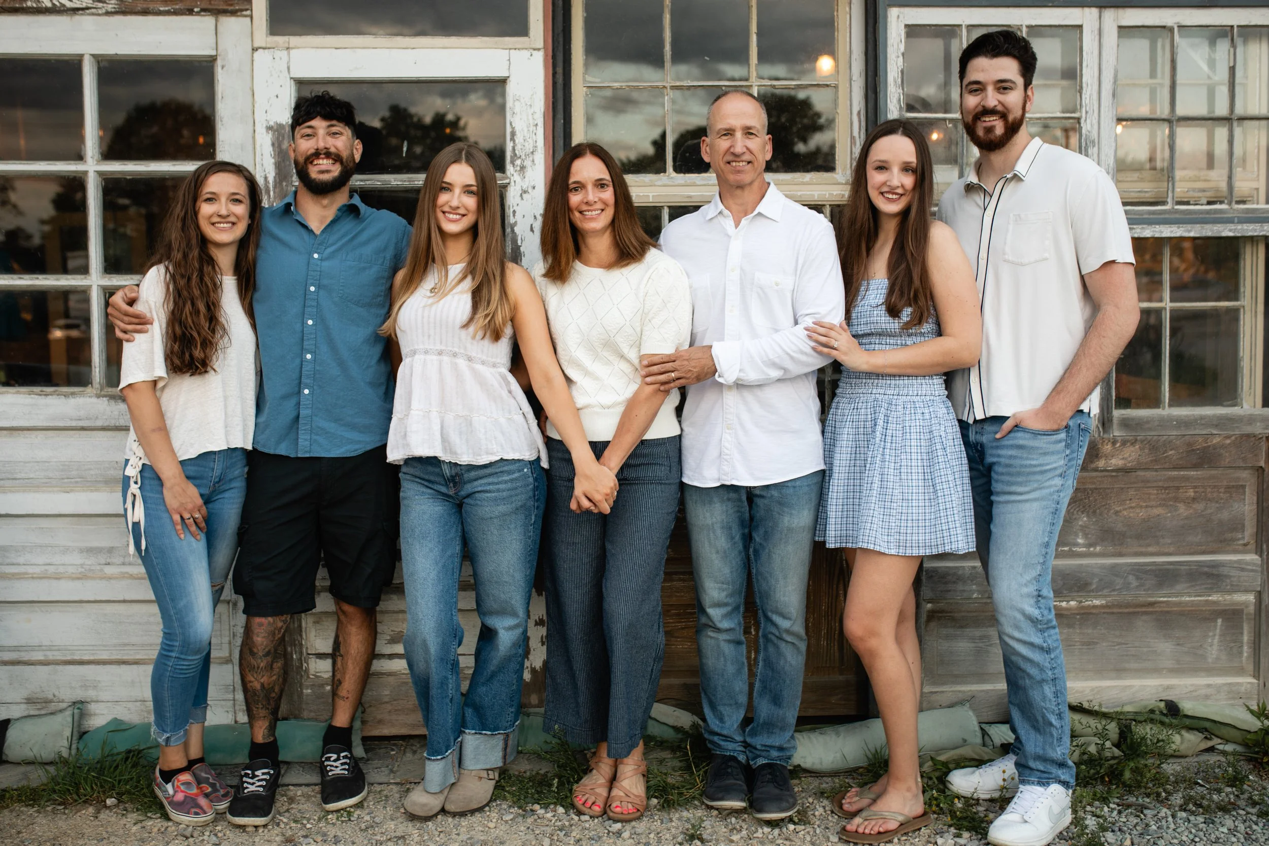 A diverse group of seven people, including men and women of different ages, standing together outdoors in front of a rustic building with large windows at sunset.