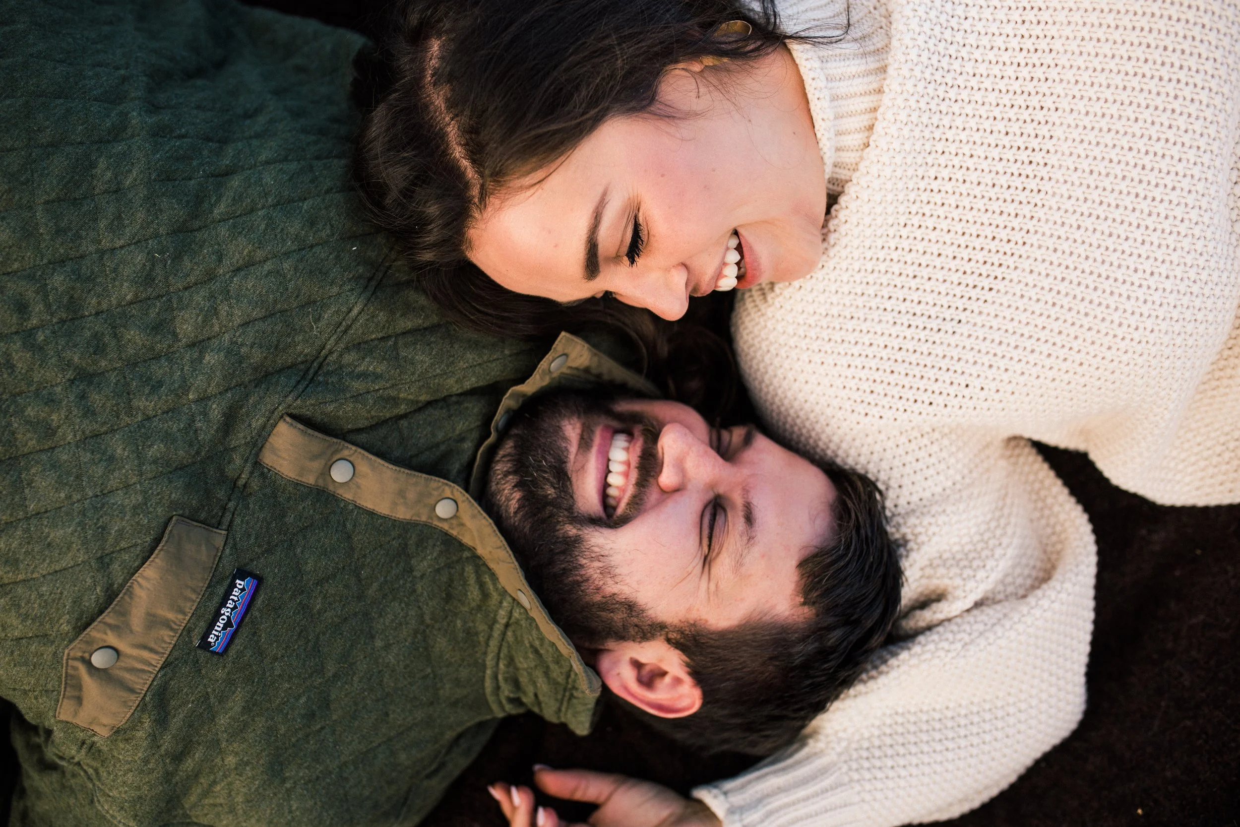 A woman and a man lying on the ground, smiling and looking at each other, close together. The woman has dark hair and is wearing a cream-colored sweater, while the man has dark hair, a beard, and is wearing a green jacket.