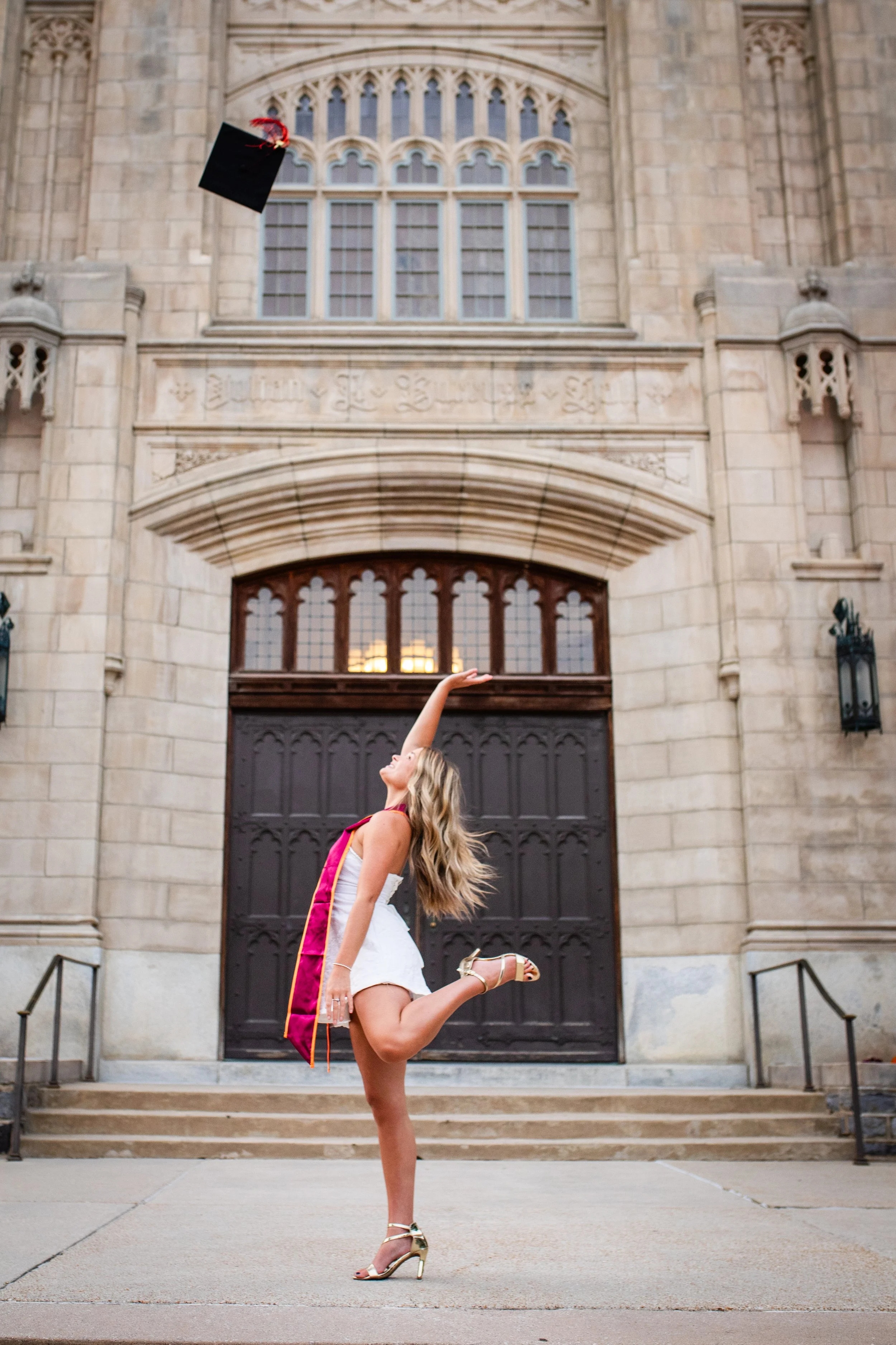 A young woman in a white dress and high heels in front of a large, historic stone building. She is wearing a graduation stole and cap, which she is tossing into the air.