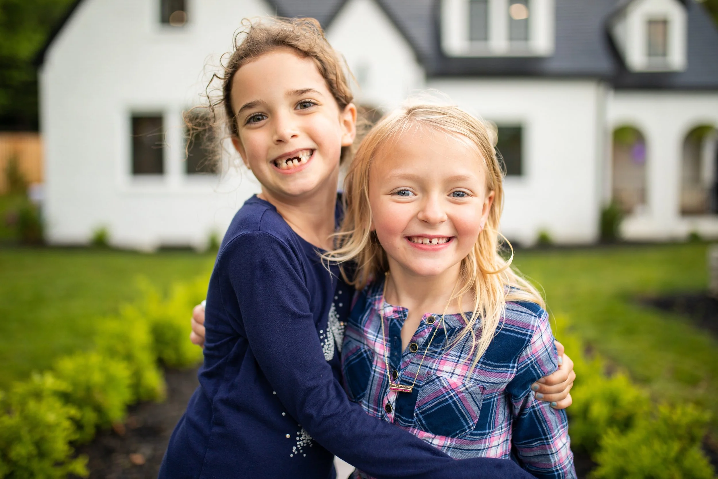 Two young girls smiling and hugging in front of a white house with green lawn and garden.
