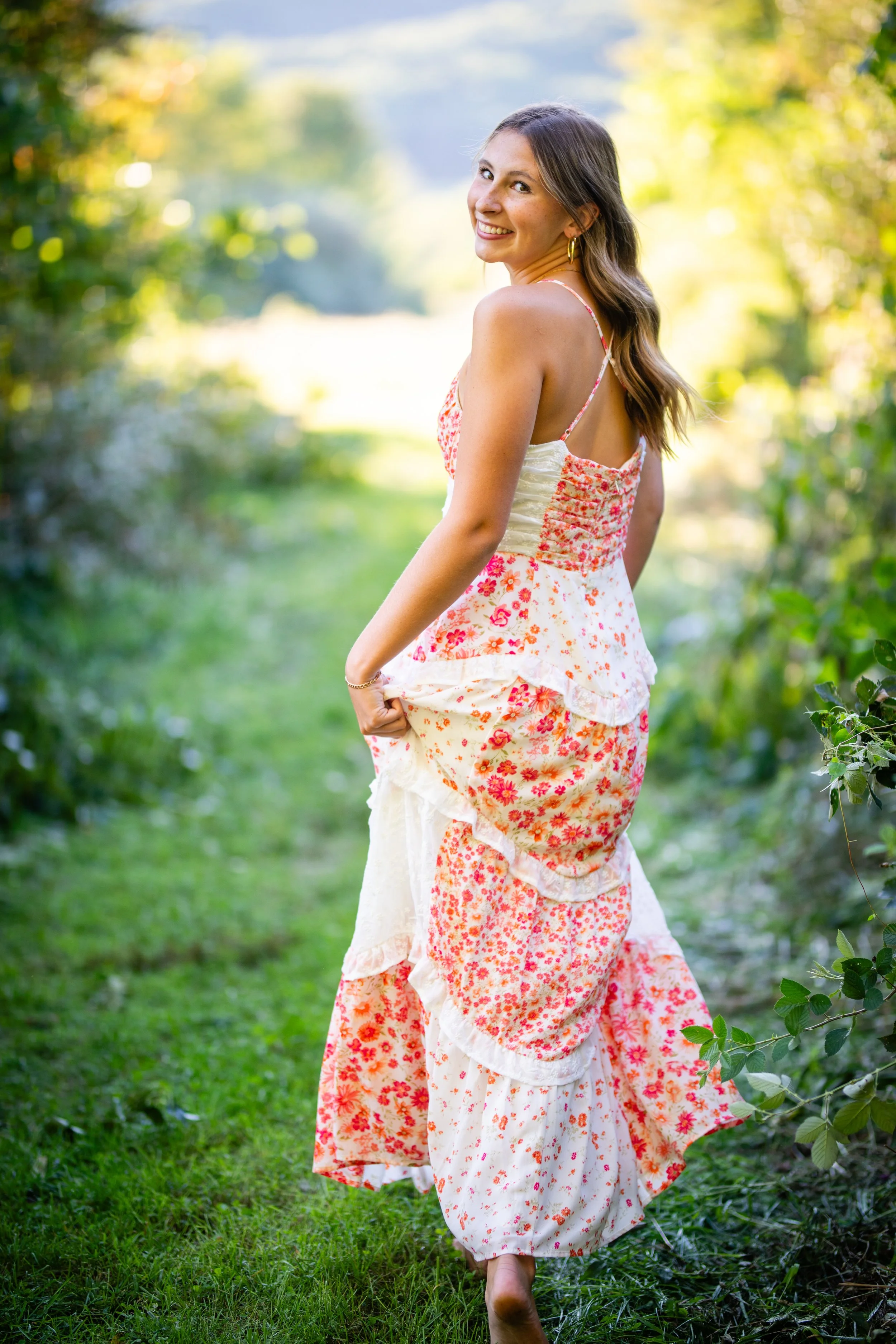 A young woman in a long, floral dress walking barefoot on a grassy path surrounded by green foliage