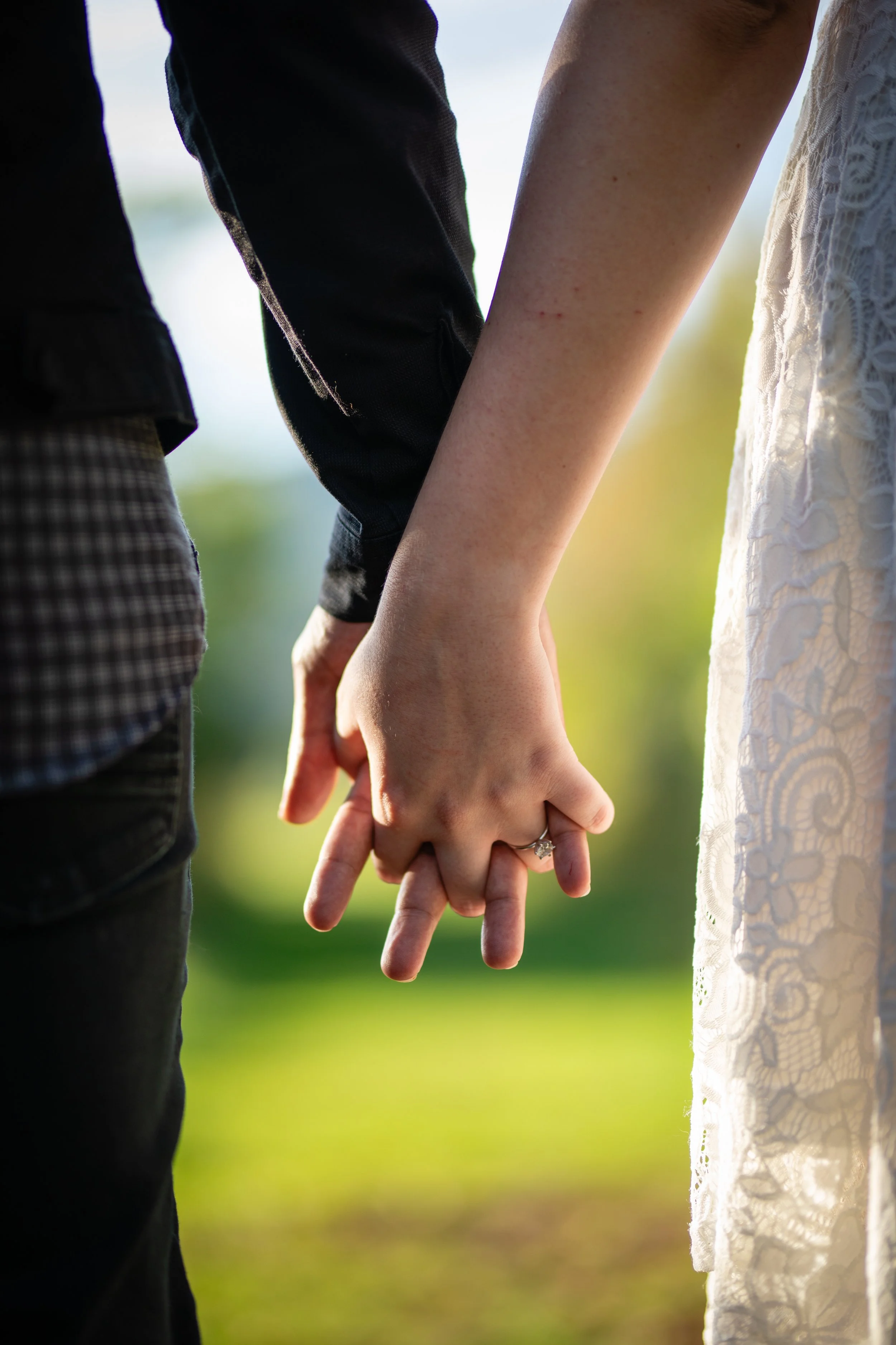 Close-up of a couple holding hands, with the woman's hand featuring an engagement ring and both people wearing casual clothing, outdoors with blurred green and yellow background.