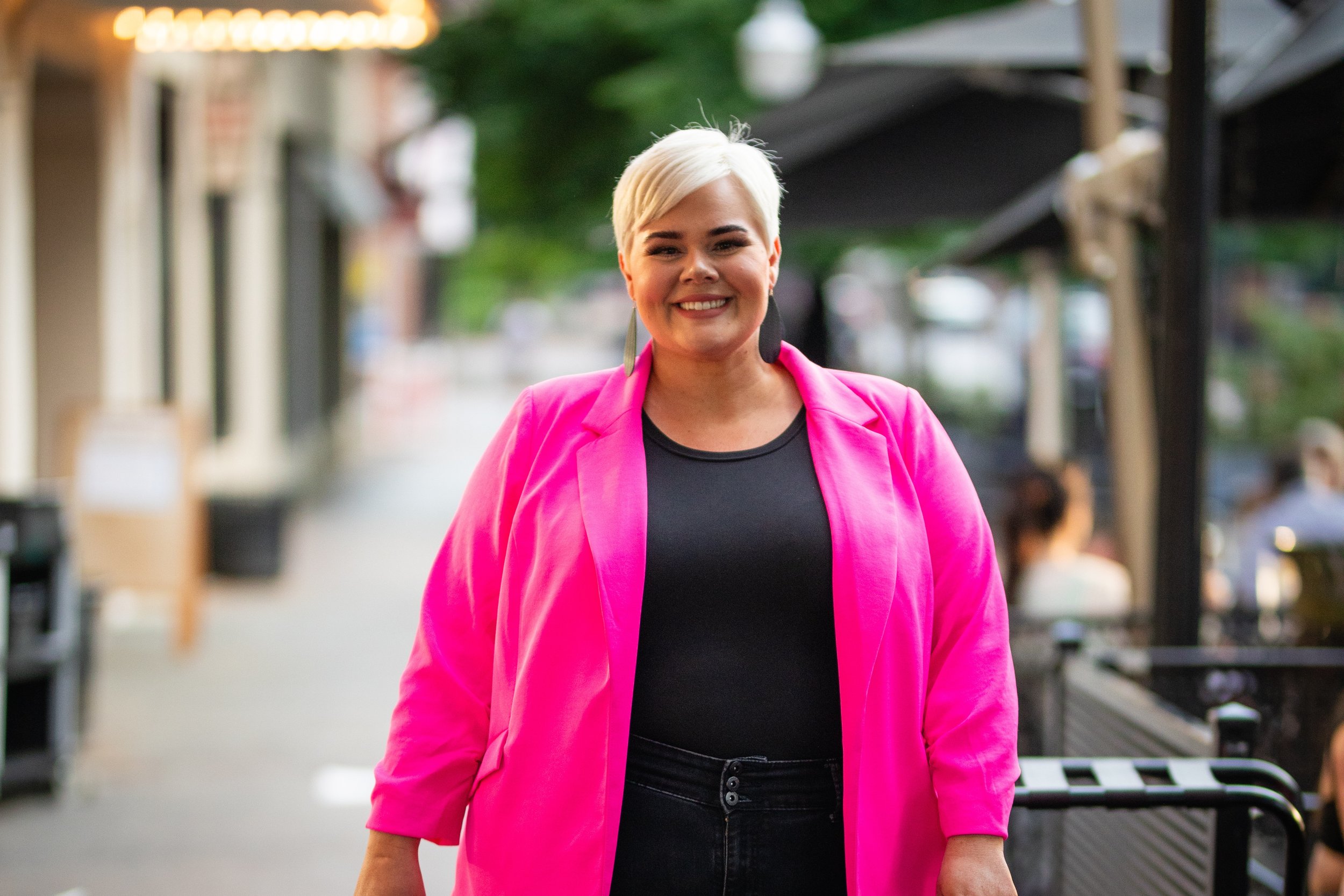 Woman smiling wearing a bright pink blazer and black top, walking outdoor on a city street.
