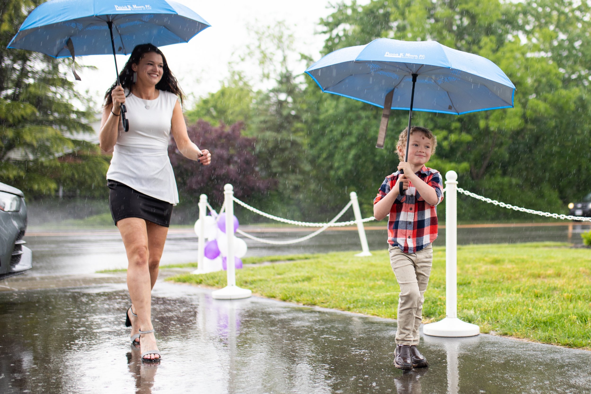 A woman and a young boy walking outdoors in the rain, holding blue umbrellas, on a wet pathway with green trees in the background.