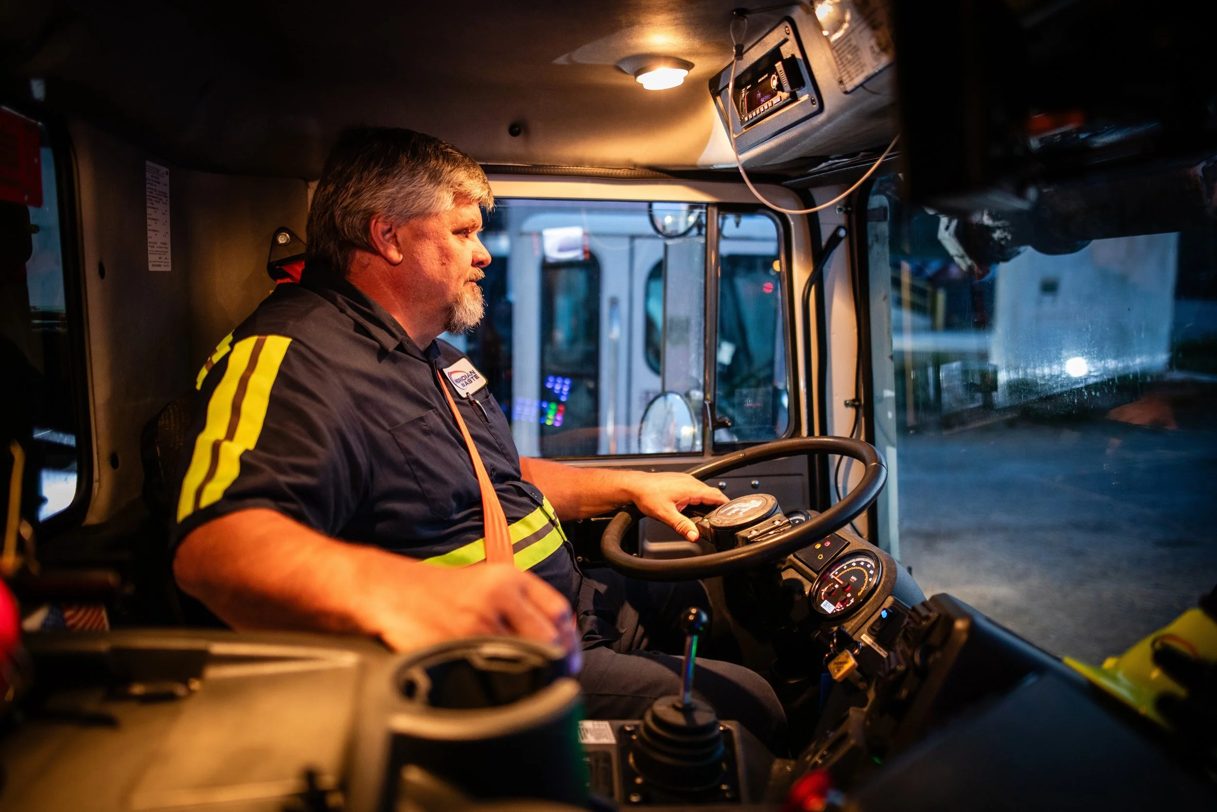 A male firefighter in uniform sitting in the driver's seat of a fire truck, looking intently at the dashboard or controls during dusk or night.