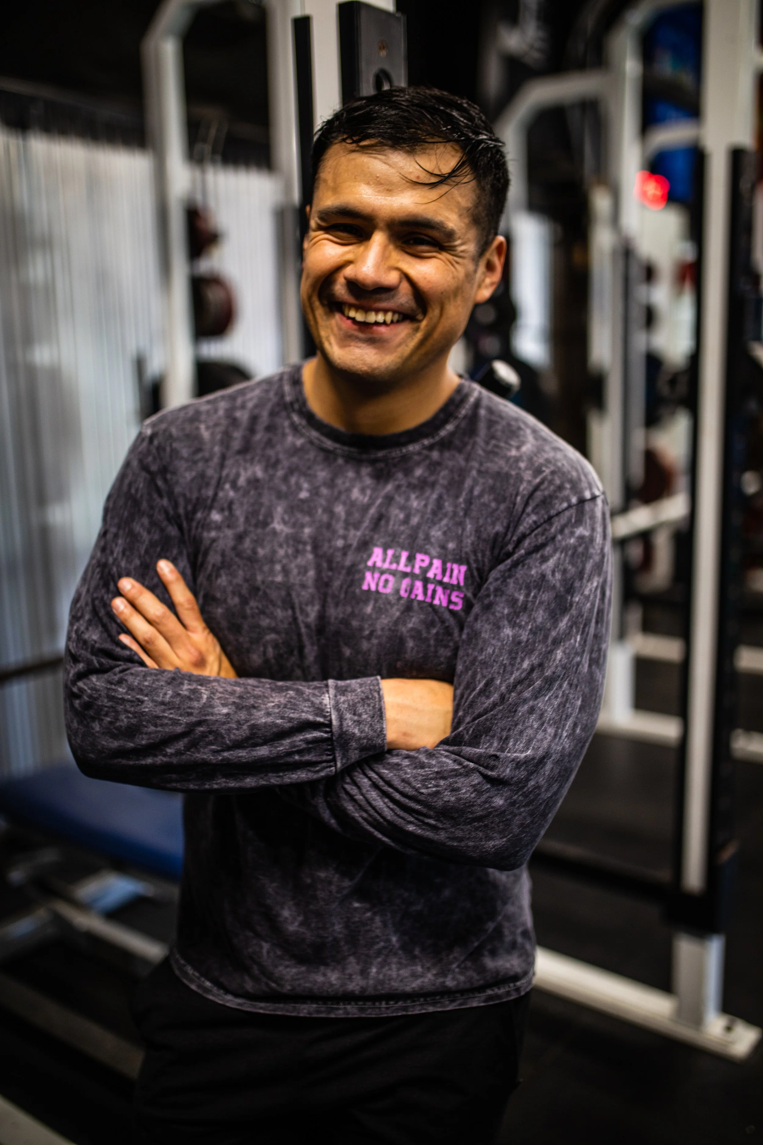 A smiling man in a gym wearing a dark, long-sleeved shirt with purple text that reads "ALL PAIN NO GAIN". He has crossed arms and is standing in front of gym equipment.