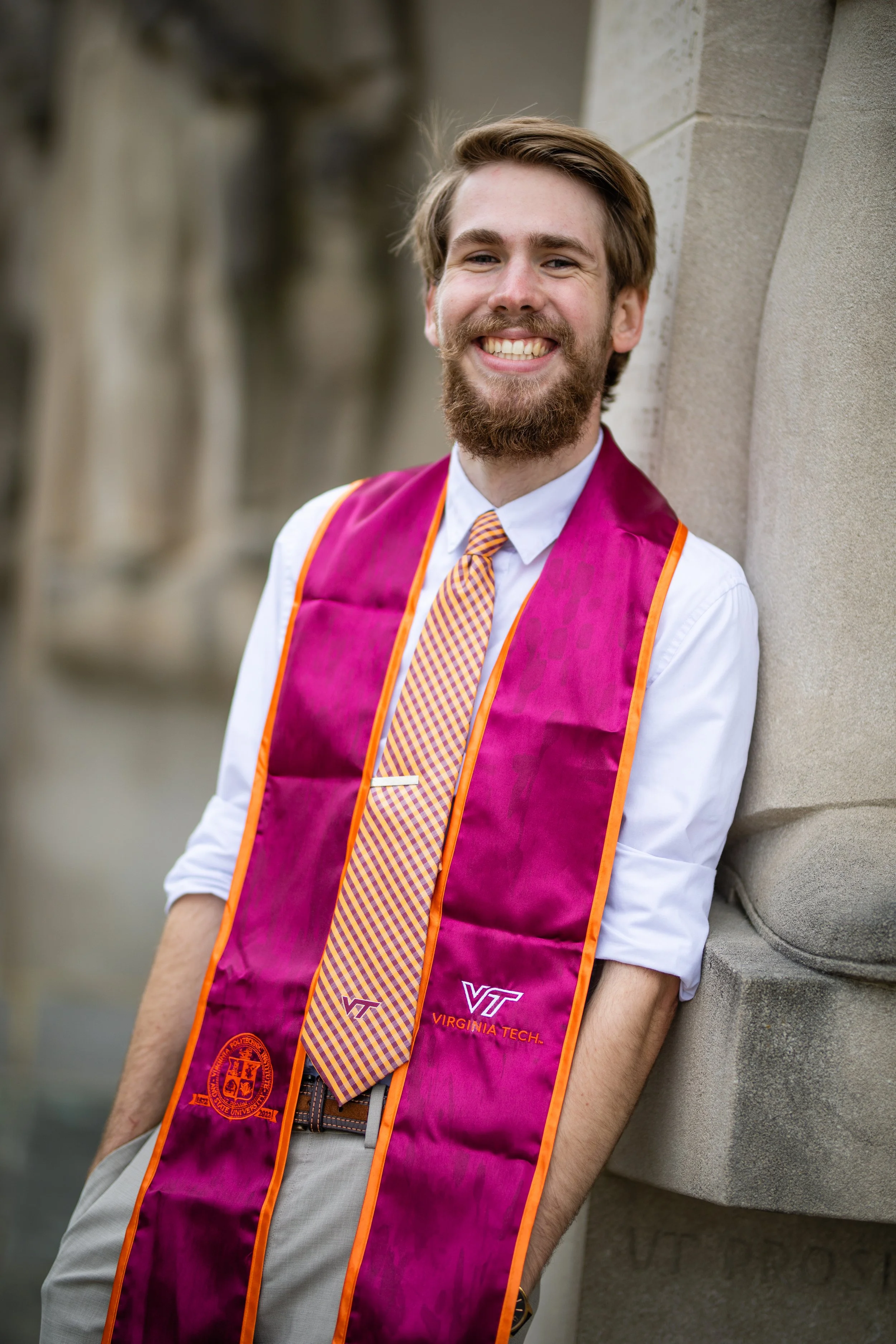 A young man with a beard and mustache smiling, wearing a white shirt, a colorful tie, and a purple graduation stole with orange edges, standing outdoors against a stone wall.