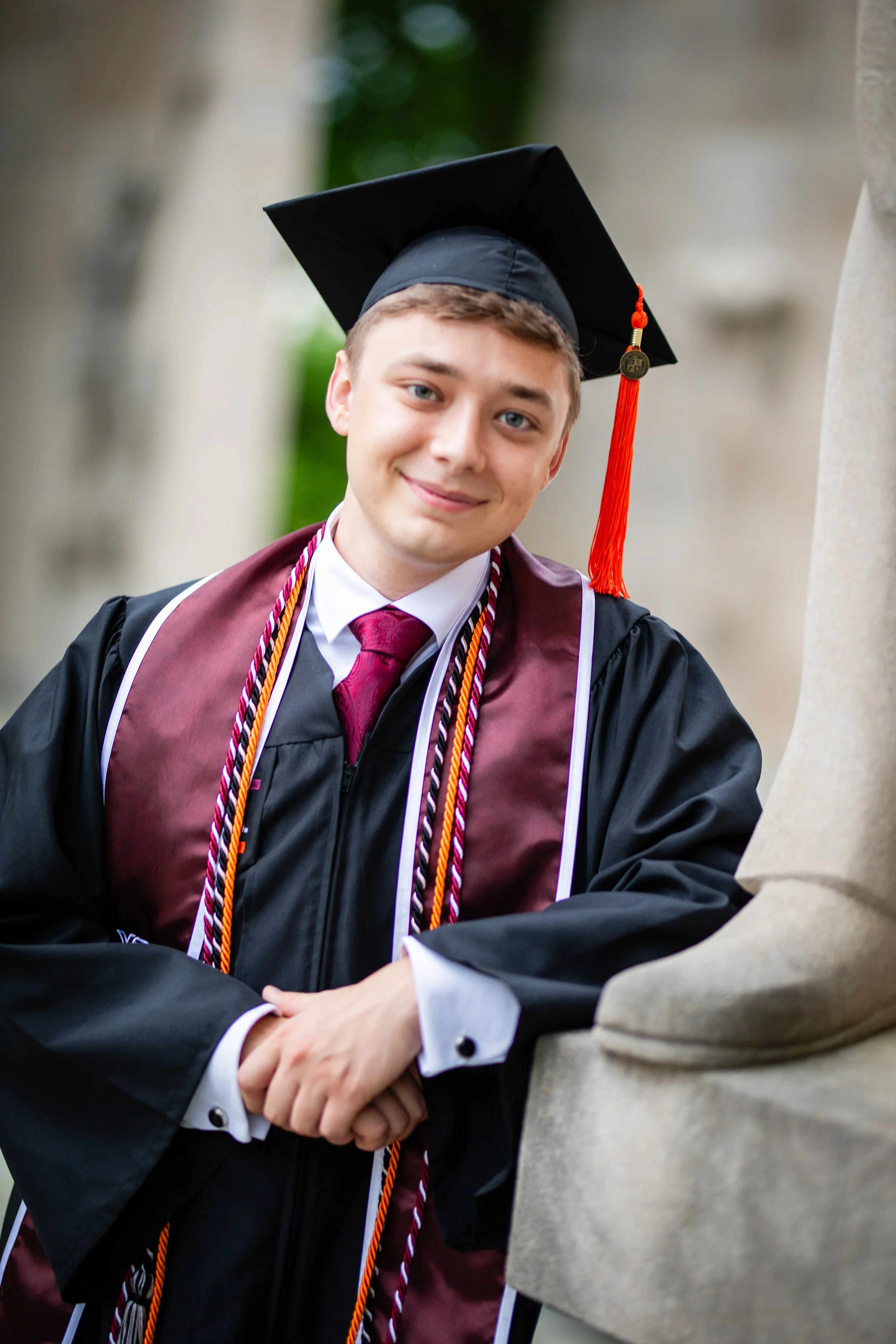 A young man in graduation gown and cap, smiling, shaking hands, outdoors with trees in the background.