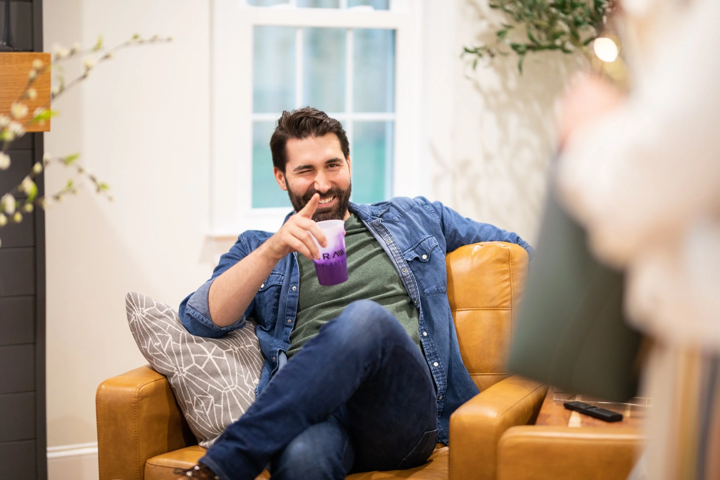 A man with dark hair and a beard sitting on a yellow leather couch, smiling and pointing his finger, holding a purple drink, in a brightly lit living room with a window in the background.