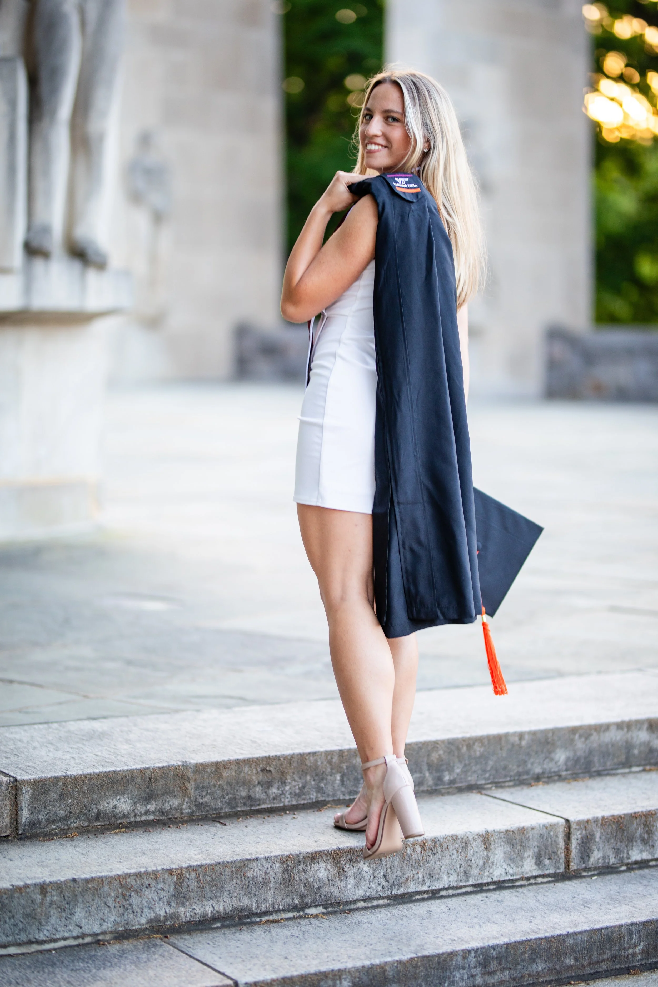 A young woman in a white dress and high heels standing on stairs with a graduation gown over her shoulder, smiling at the camera.