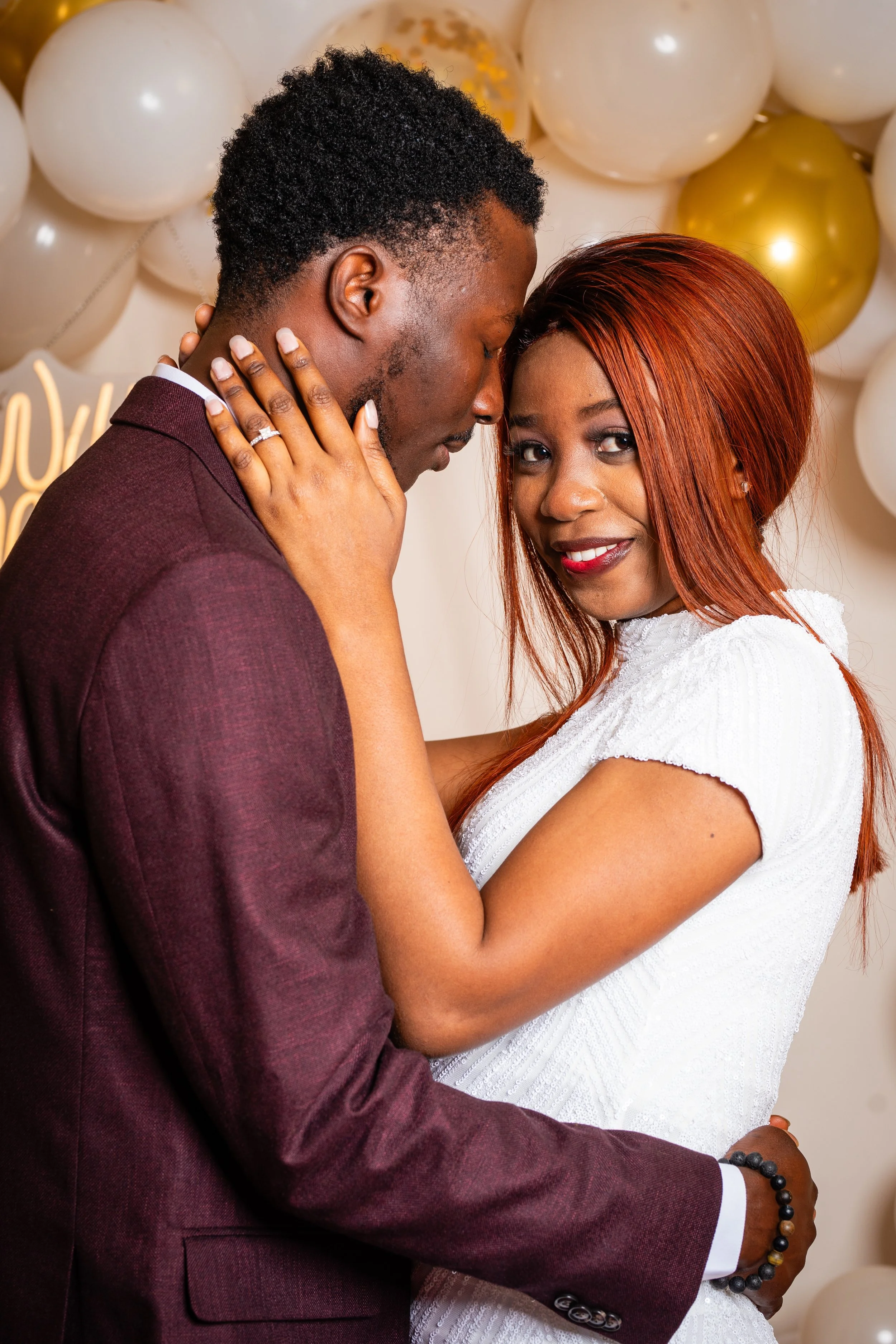 A couple sharing an intimate moment at a celebration, with balloons and a 'Love You' sign in the background.