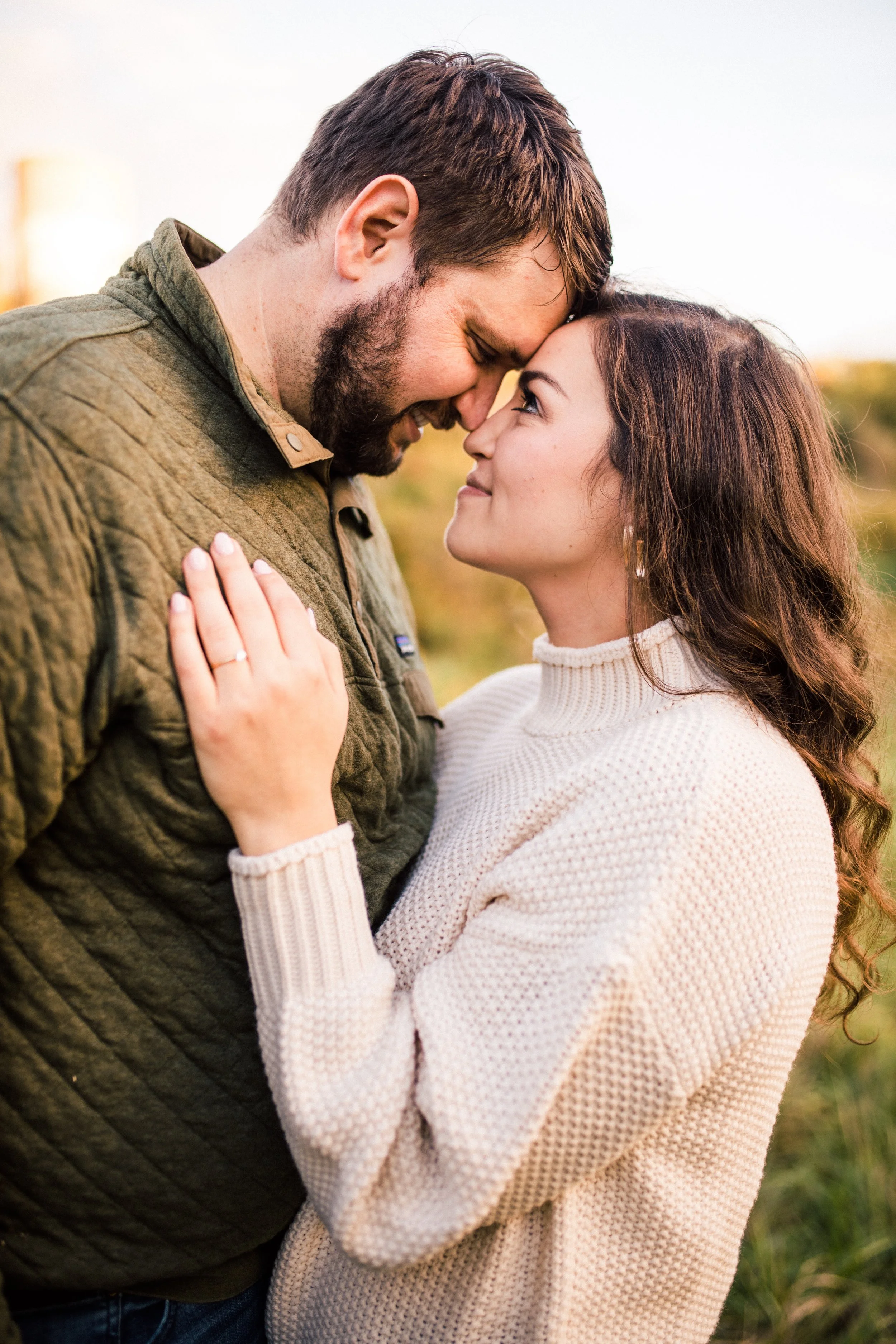 A couple standing close, smiling, with their foreheads touching, outdoors during sunset.