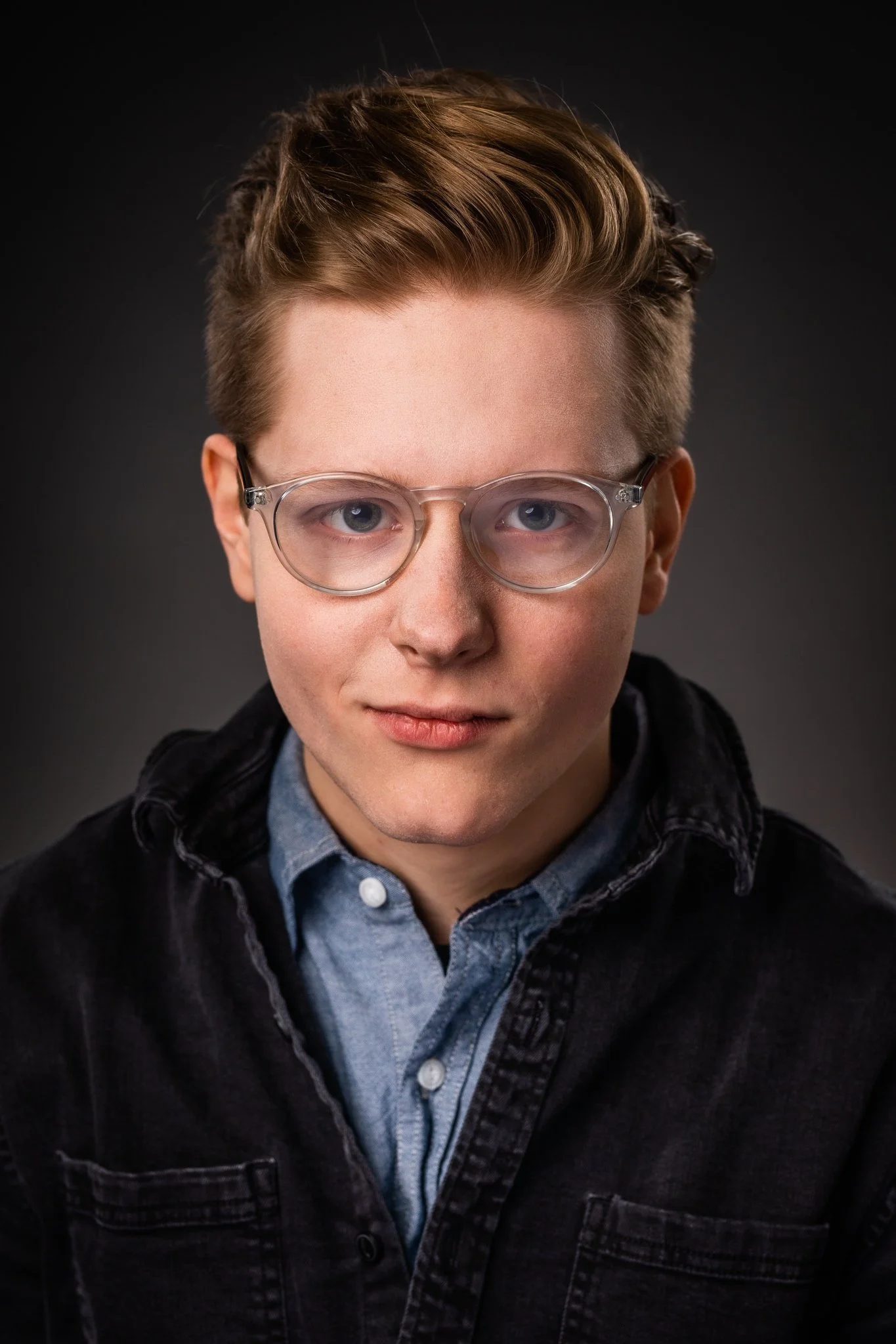 A young man with light brown hair, wearing clear glasses, a blue button-up shirt, and a black jacket, looking directly at the camera against a dark background.