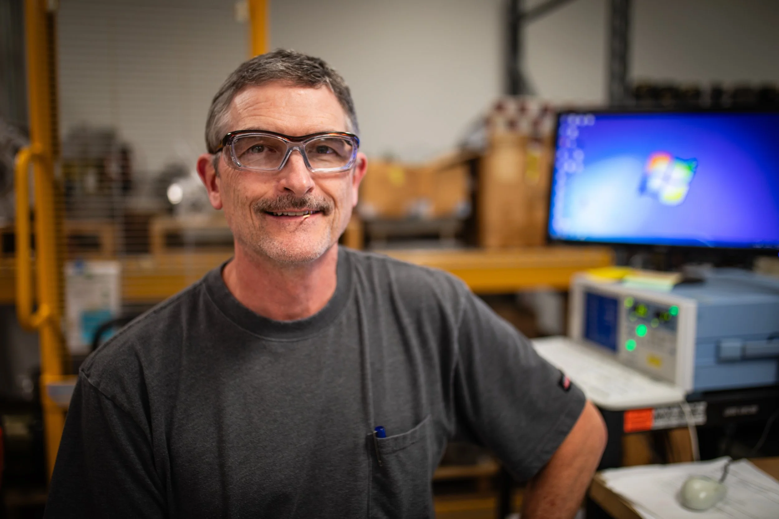 A smiling man wearing safety glasses sits in a workshop with a computer and electronic equipment in the background.