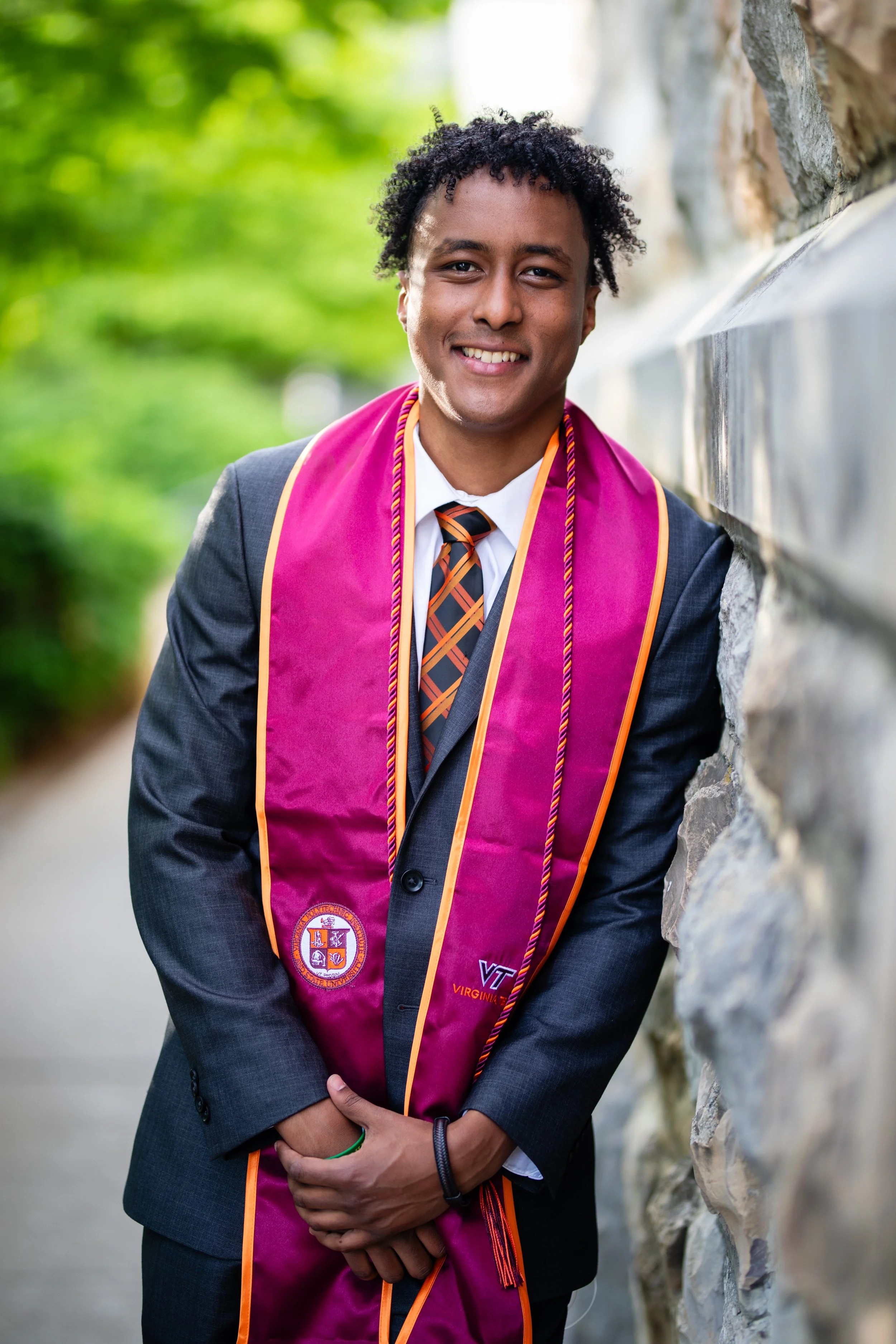 A young man in a gray suit with a Virginia Tech graduation stole, smiling, standing outdoors near a stone wall, with green foliage in the background.