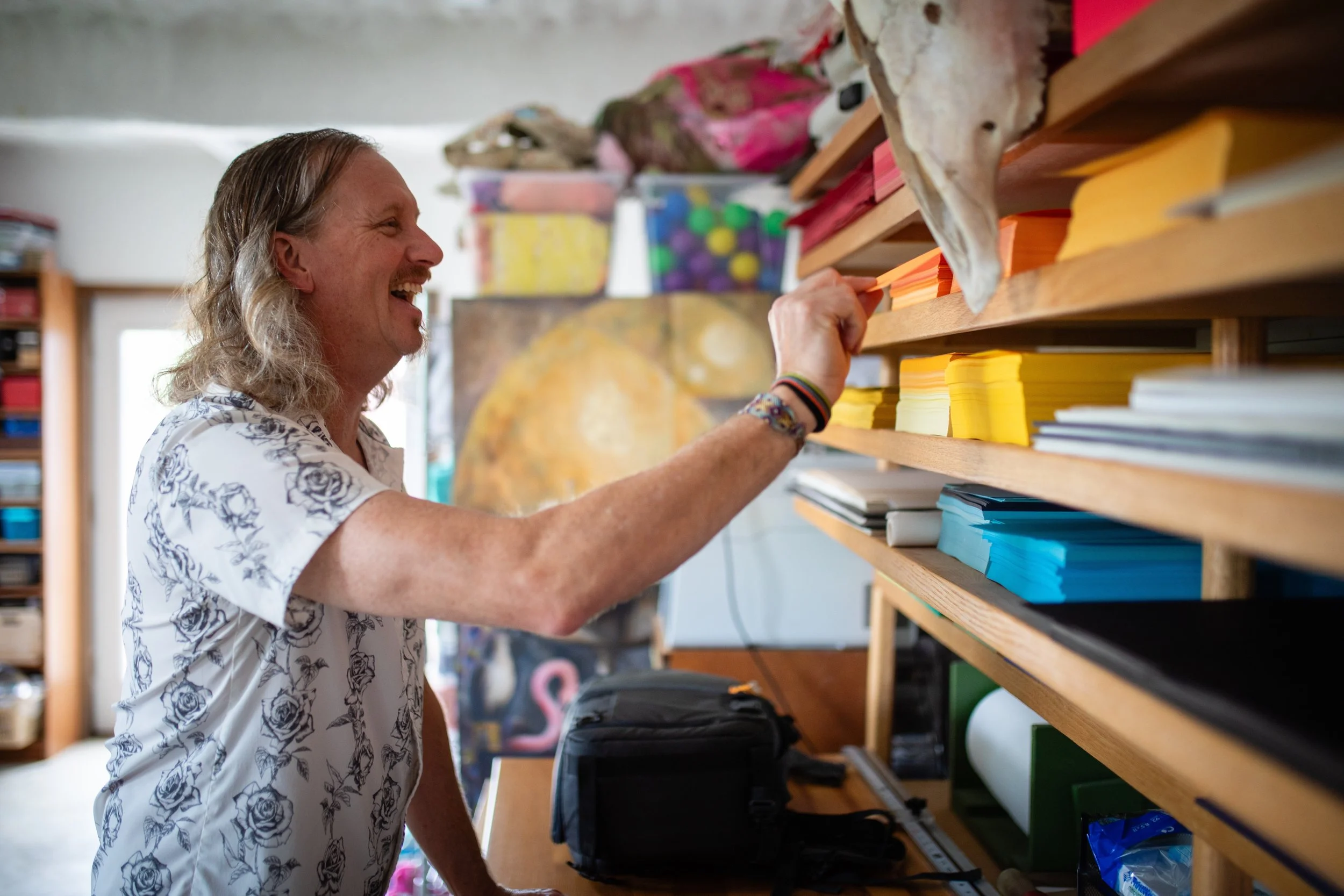 A man with long gray hair, wearing a white shirt with blue roses, is smiling and reaching for yellow sticky notes on a wooden shelf in an art or craft room.